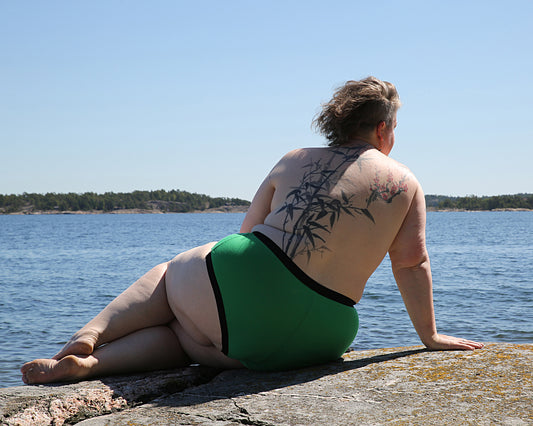 Back view of a model wearing The Bamboo Hipster - Limited Edition in colour Green. The model is sitting in a relaxed pose on a cliff, looking out over the sea on a summer day.