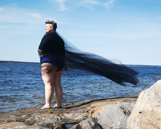 Back side view of a model wearing a pair of Bamboo Brazilians in the blue colourway. The model is standing on a rocky cliff, looking out over the sea. She has tulle draped around her shoulders and it's billowing and flowing on a breeze.
