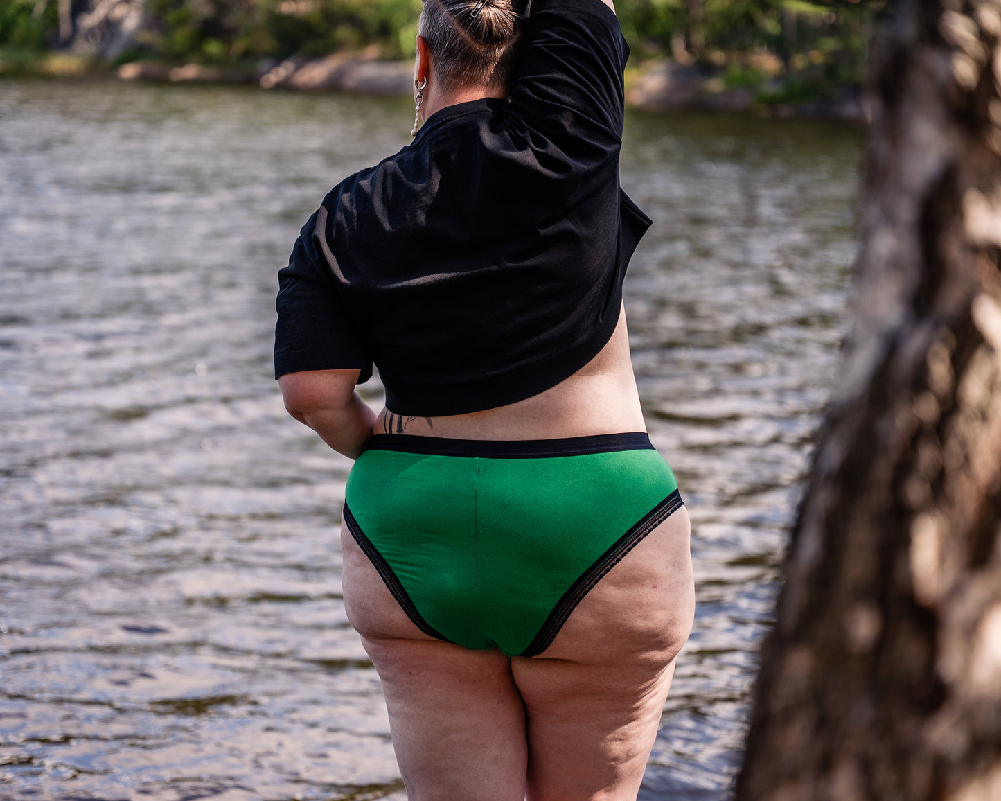 Back view of a model wearing a pair of The Bamboo High Cut knickers in the green colourway, with a black t-shirt. She is standing by a lake, with a blurry tree trunk in the foreground.