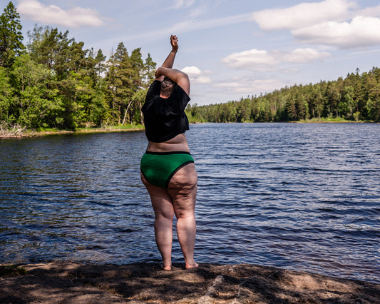 Back side view of a model wearing a pair of The Bamboo High Cut in the green colourway, with a black t-shirt. She is standing on a rockcky outcroop by a lake, with trees in the background.