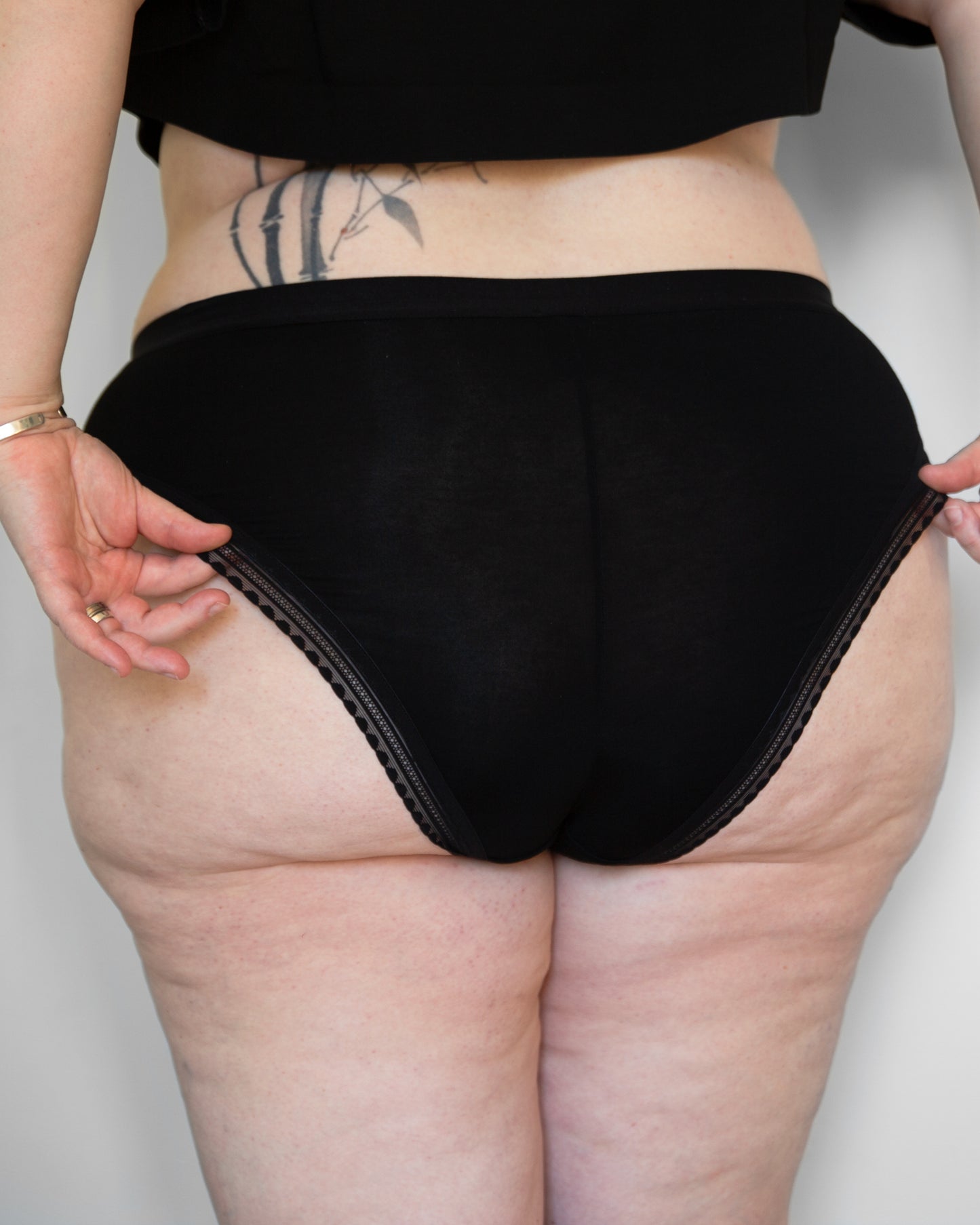 Back view of a model wearing The Bamboo High Cut in black colour with standard elastic in the leg opening. The models’ hands are adjusting the top leg opening on both sides.