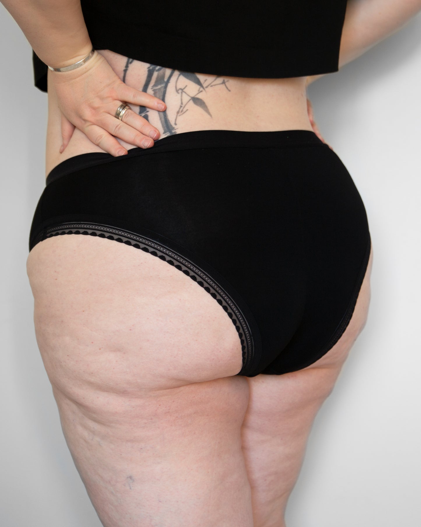 Back side view of a model wearing The Bamboo High Cut in black colour with standard elastic in the leg opening. The models’ hands are resting on her lower back.
