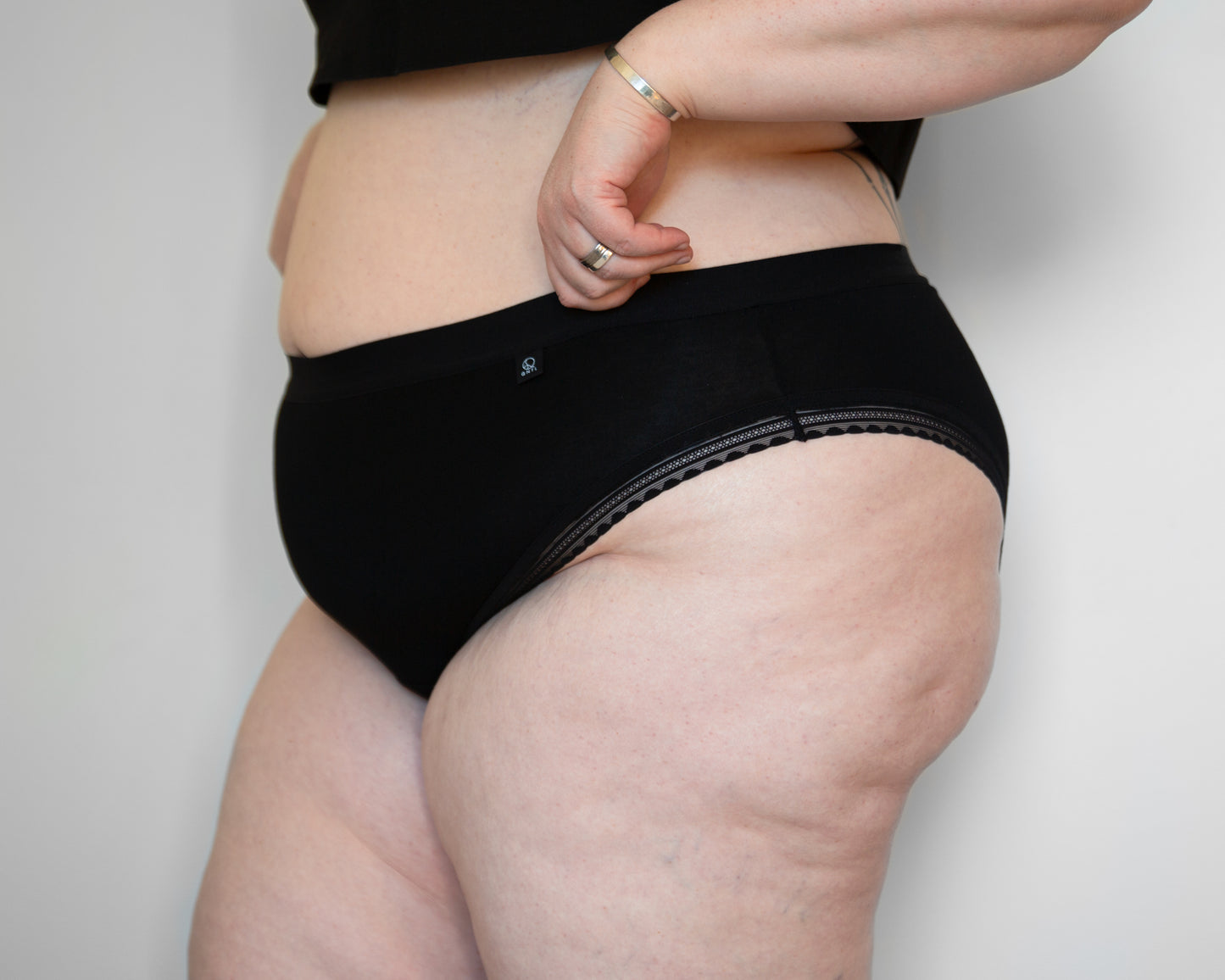 Front side view of a model wearing The Bamboo High Cut in black colour with standard elastic in the leg opening. The models’ hands are holding the edge of the waistband, just above the brand label.