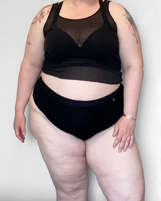 Front view of a model wearing The Bamboo Thong in the black colourway, with a sheer top and bra. She is standing in a relaxed pose, with one hand on her thigh, infront of a light background.