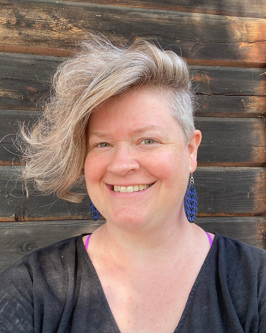 Model wearing blue and black coloured oval earrings. She is smiling and sitting infront of a dark wooden wall.