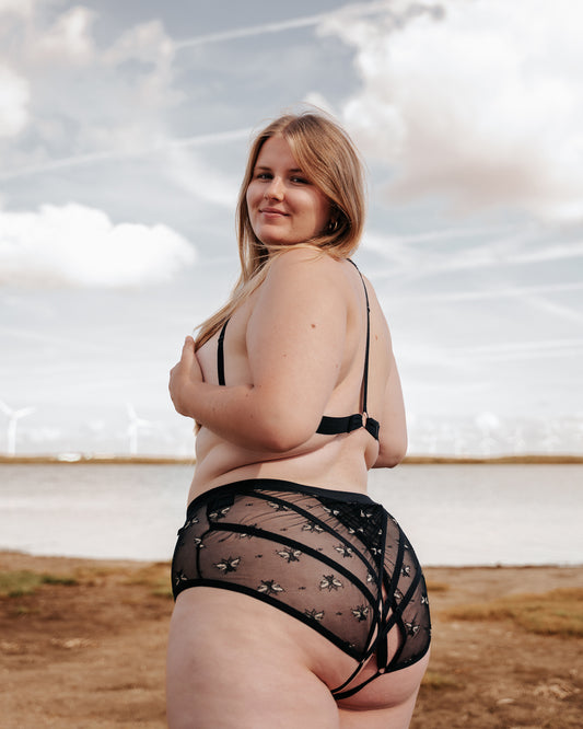 Side view of a model wearing The Cage Bra and The Crossover Hipster, walking forwards the water on the beach. Model is looking over her shoulder towards the viewer, and you can see the single back strap of The Cage Bra.