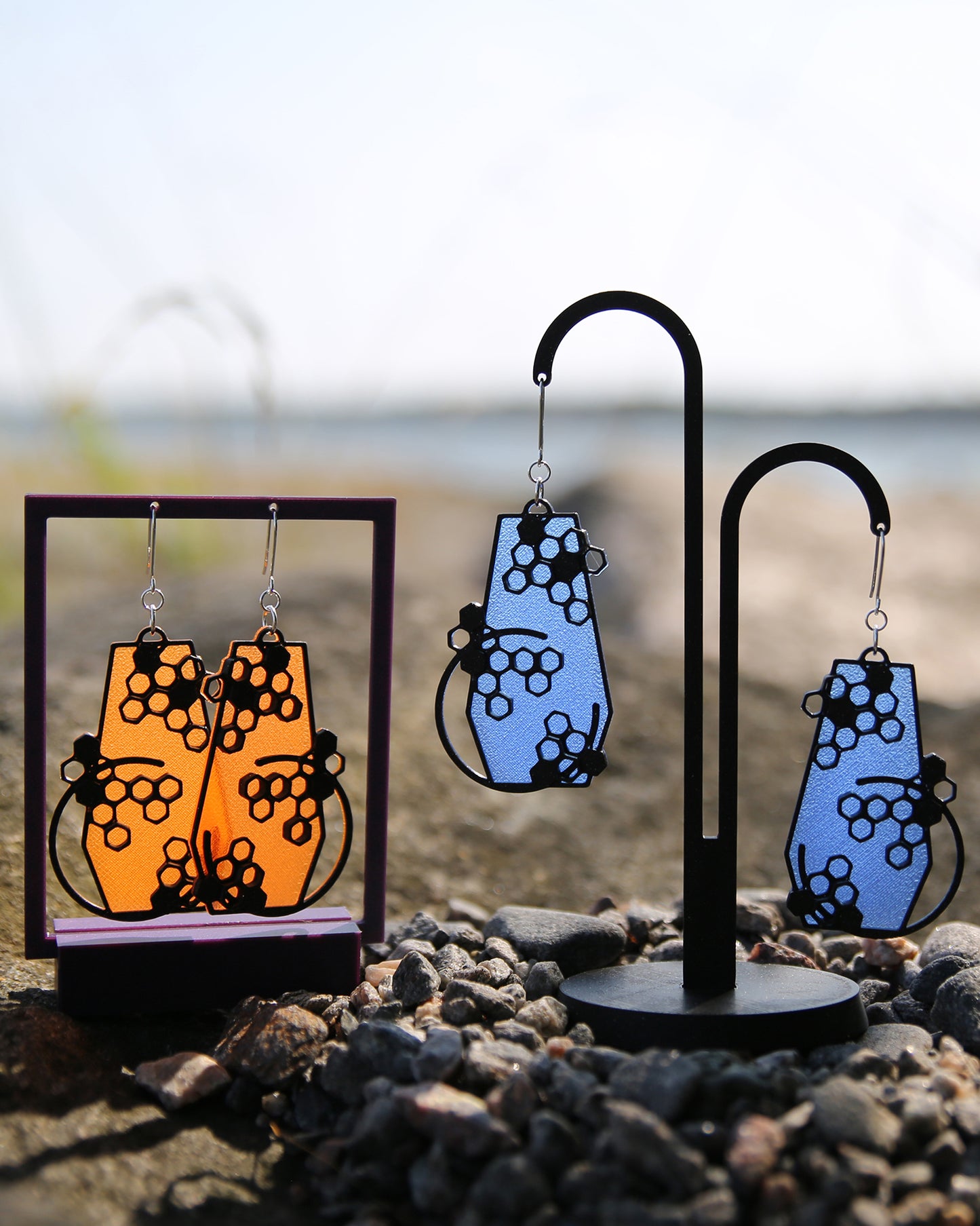 Two pairs of earrings, orange to the left and blue to the right, haning on stands with a blurred natural background. The sun is shining through the translucent bottom of the earrings.
