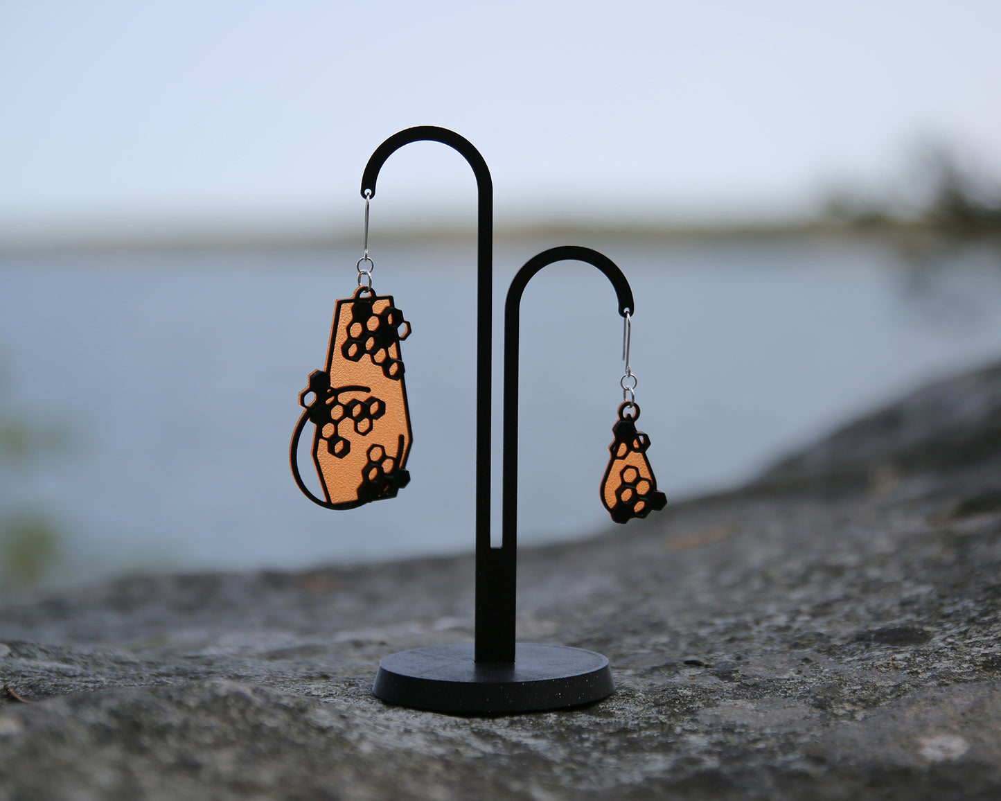 Two earrigs with a honeycomb design, displayed on a black stand with a blurred natural background. The earring to the left is the original big size and the earring to the right is a small verion of the original design. They are both orange and black.