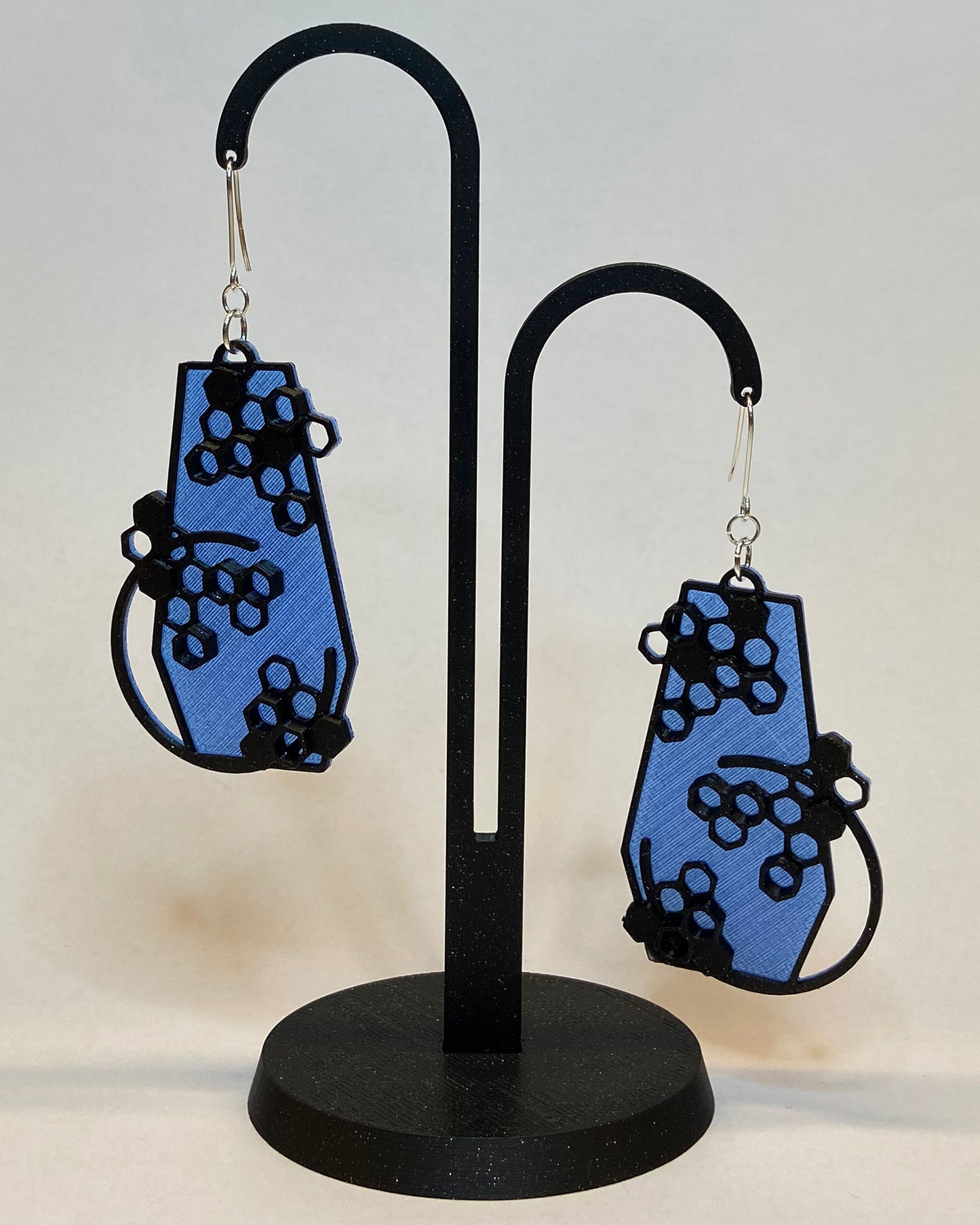 A pair of 3D-printed earrings with a honeycomb design on a black stand against a light background. The earrings has two colours, a blue coloured bottom and contrasting black pattern. The fish hook is made of silver.