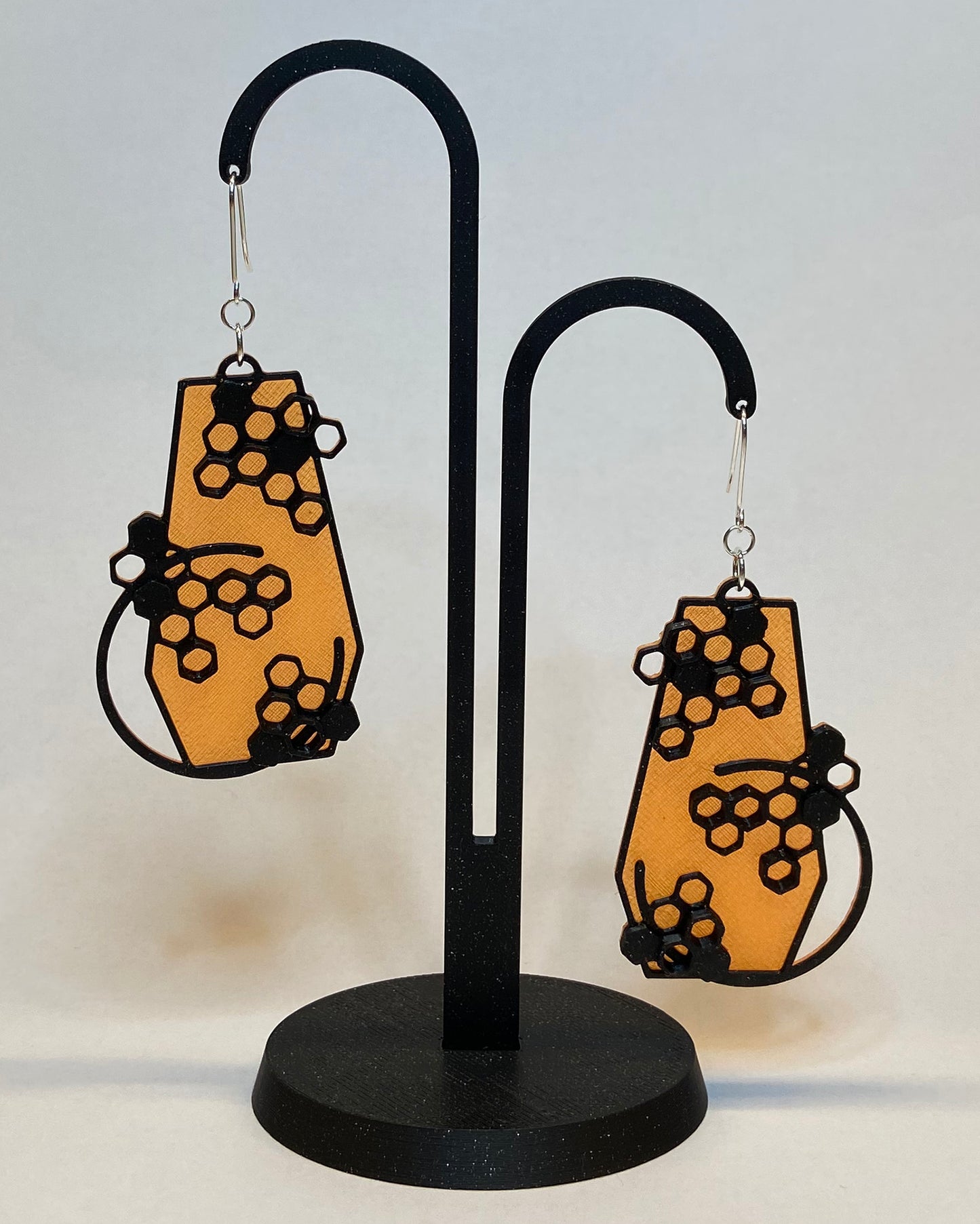 A pair of 3D-printed earrings with a honeycomb design on a black stand against a light background. The earrings has two colours, an orange coloured bottom and contrasting black pattern. The fish hook is made of silver.