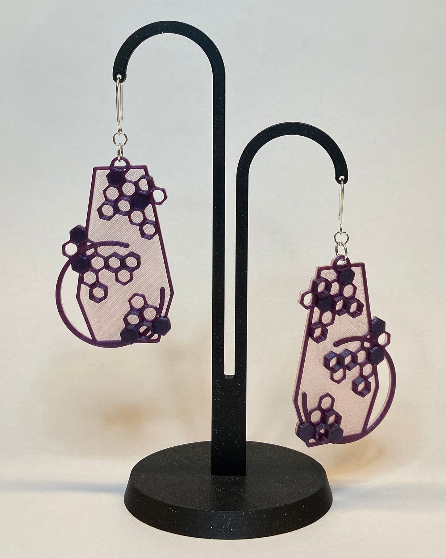 A pair of 3D-printed earrings with a honeycomb design on a black stand against a light background. The earrings has two colours, a pink coloured bottom and contrasting purple pattern. The fish hook is made of silver.