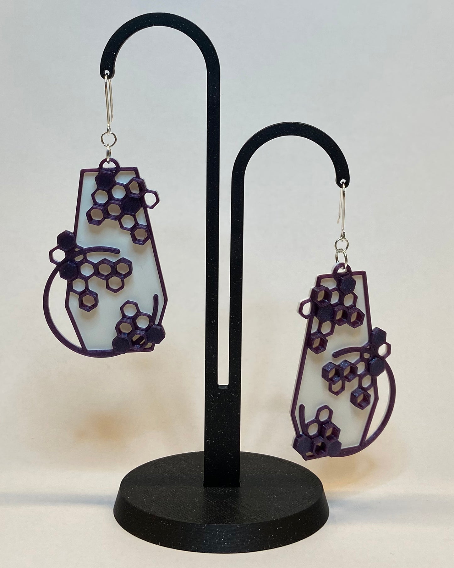 A pair of 3D-printed earrings with a honeycomb design on a black stand against a light background. The earrings has two colours, a white coloured bottom and contrasting purple pattern. The fish hook is made of silver.