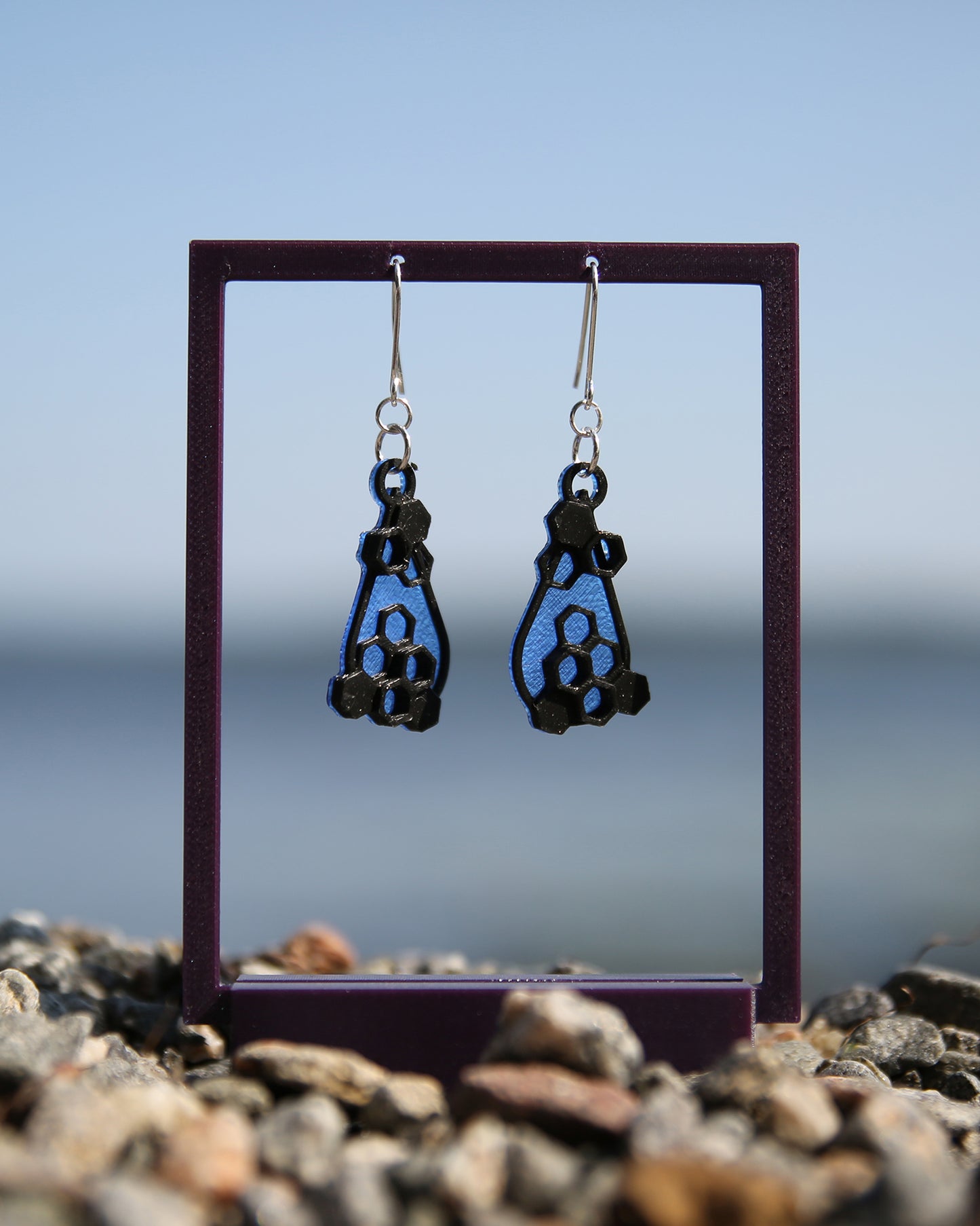A pair of blue and black earrings with a honeycomb design, haning on a purple stand with a blurred natural background.