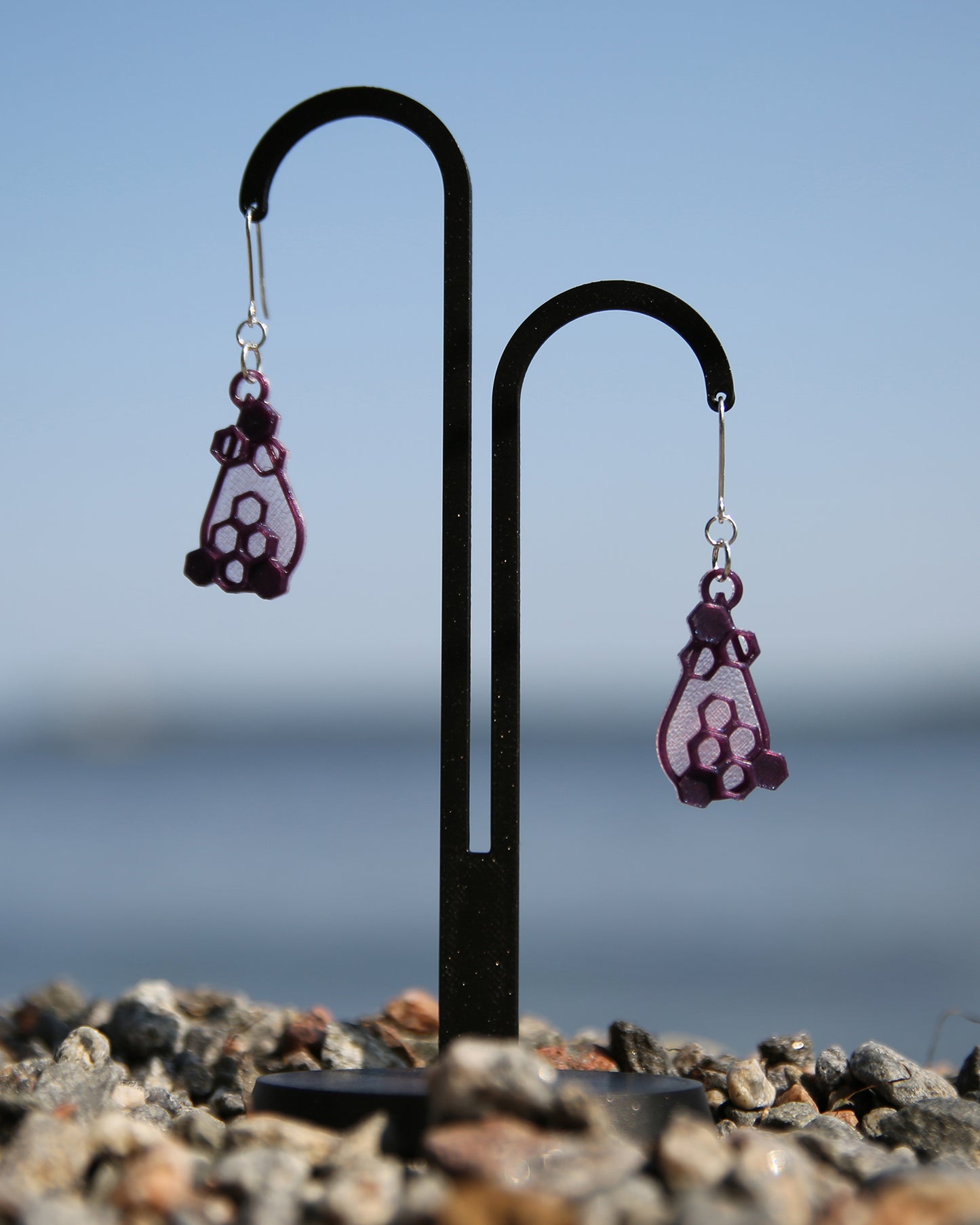 A pair of light pink and purple earrings with a honeycomb design, haning on a black stand with a blurred natural background.