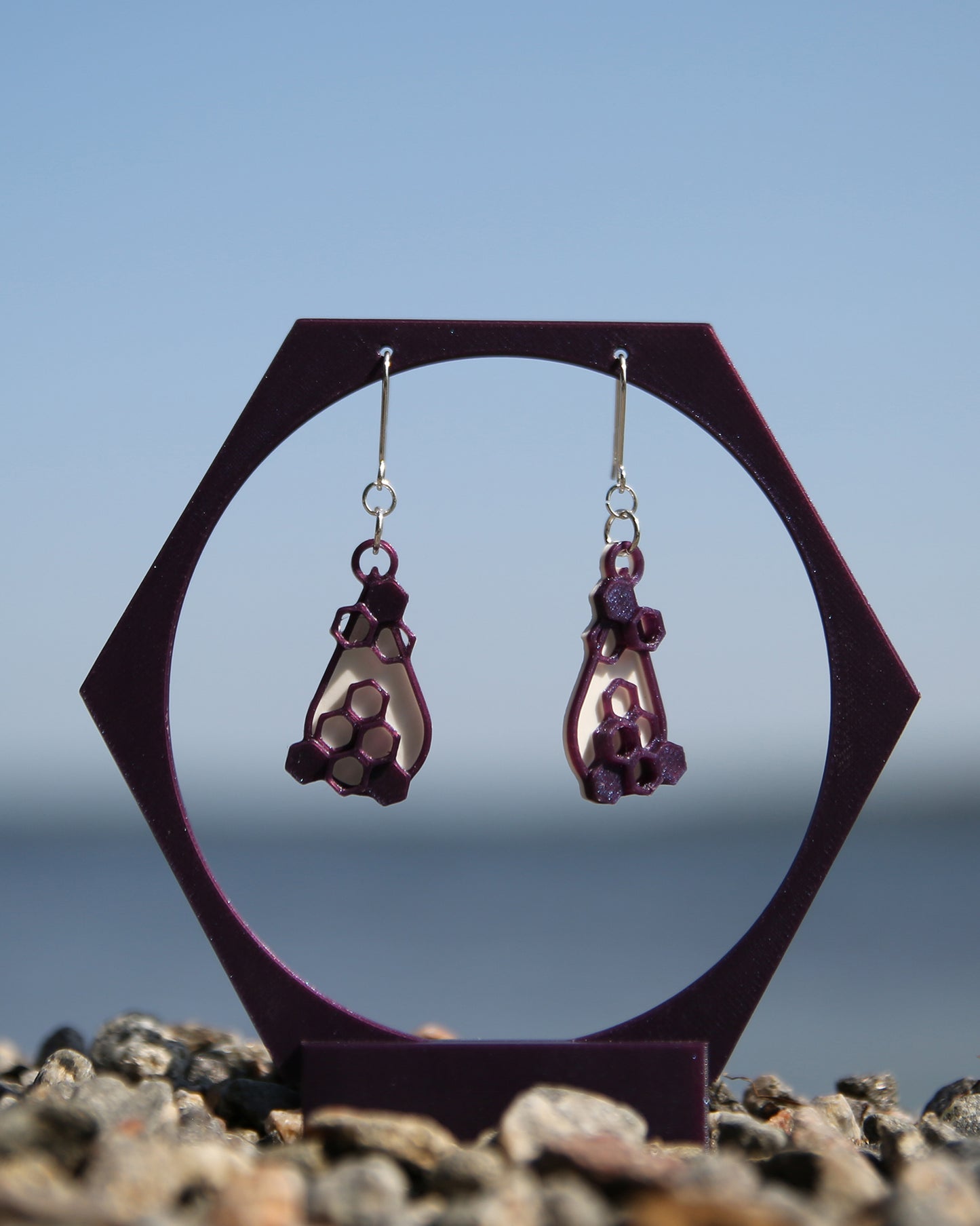 A pair of white and purple earrings with a honeycomb design, haning on a purple stand with a blurred natural background.