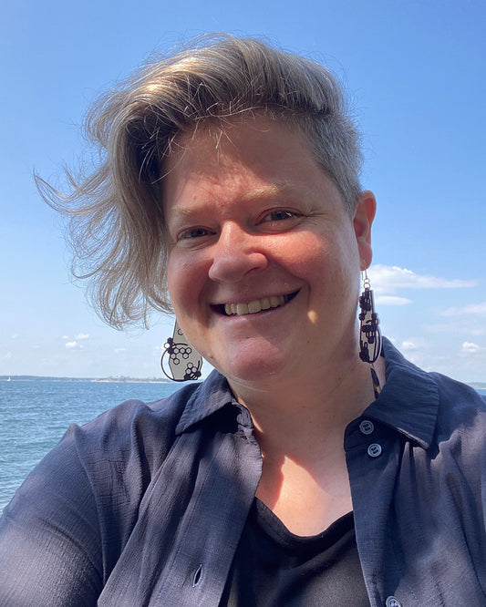 A model is sitting infront of the sea, wearing a pair of geometrical shaped earrings with a honeycomb design. The earrings are white and purple in colour.