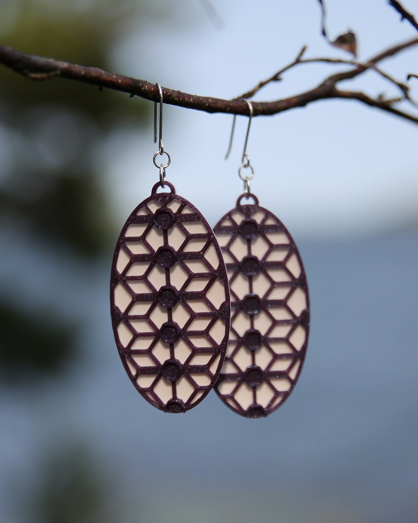 A pair of oval shaped earrings with a lace inspired design, hanging on a branch. The earrings have a white coloured background, and the pattern has a glittering purple colour.
