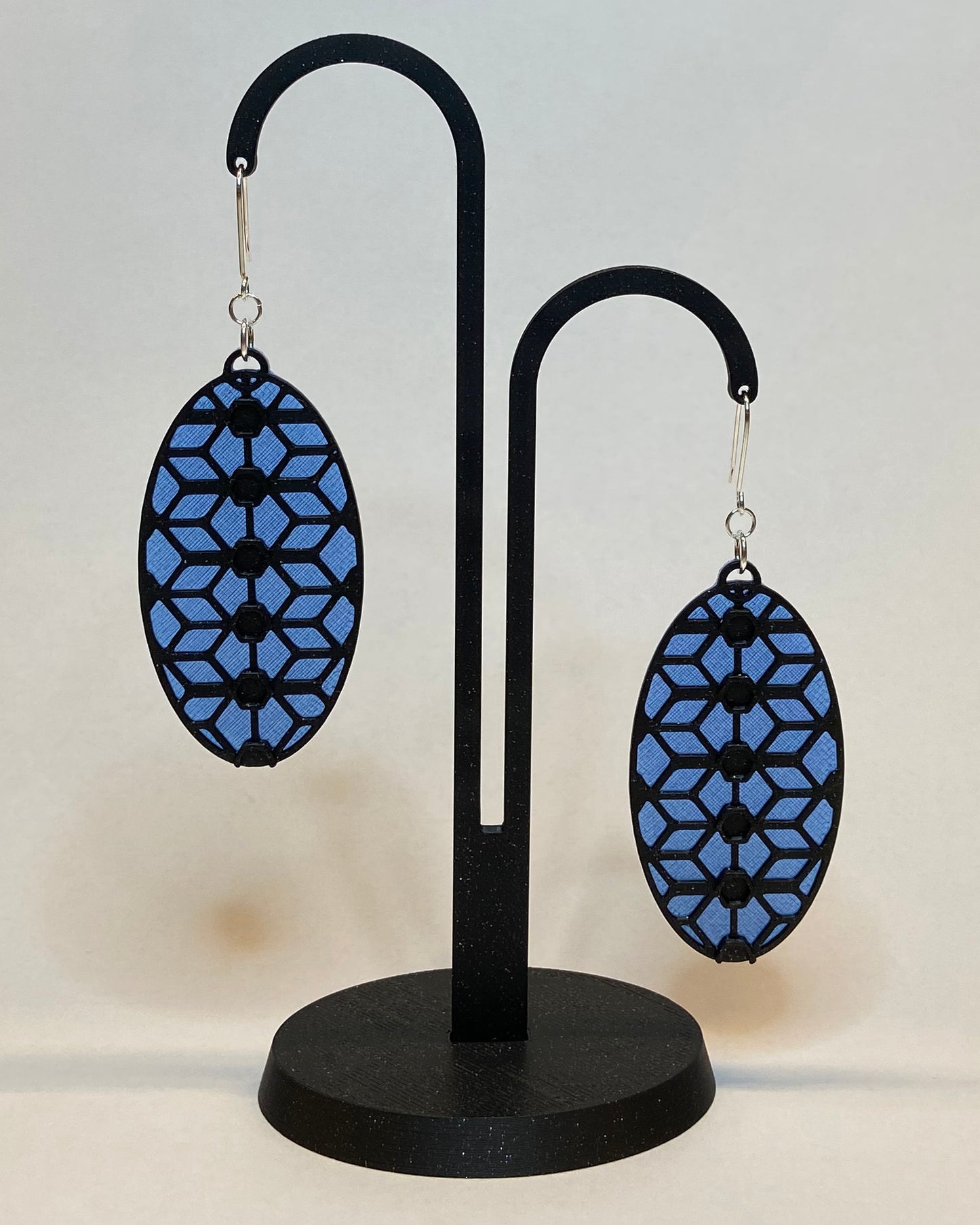 A pair of 3D-printed earrings with a lace inspiered design on a black stand against a light background. The earrings has two colours, a blue coloured bottom and contrasting black pattern. The fish hook is made of silver.