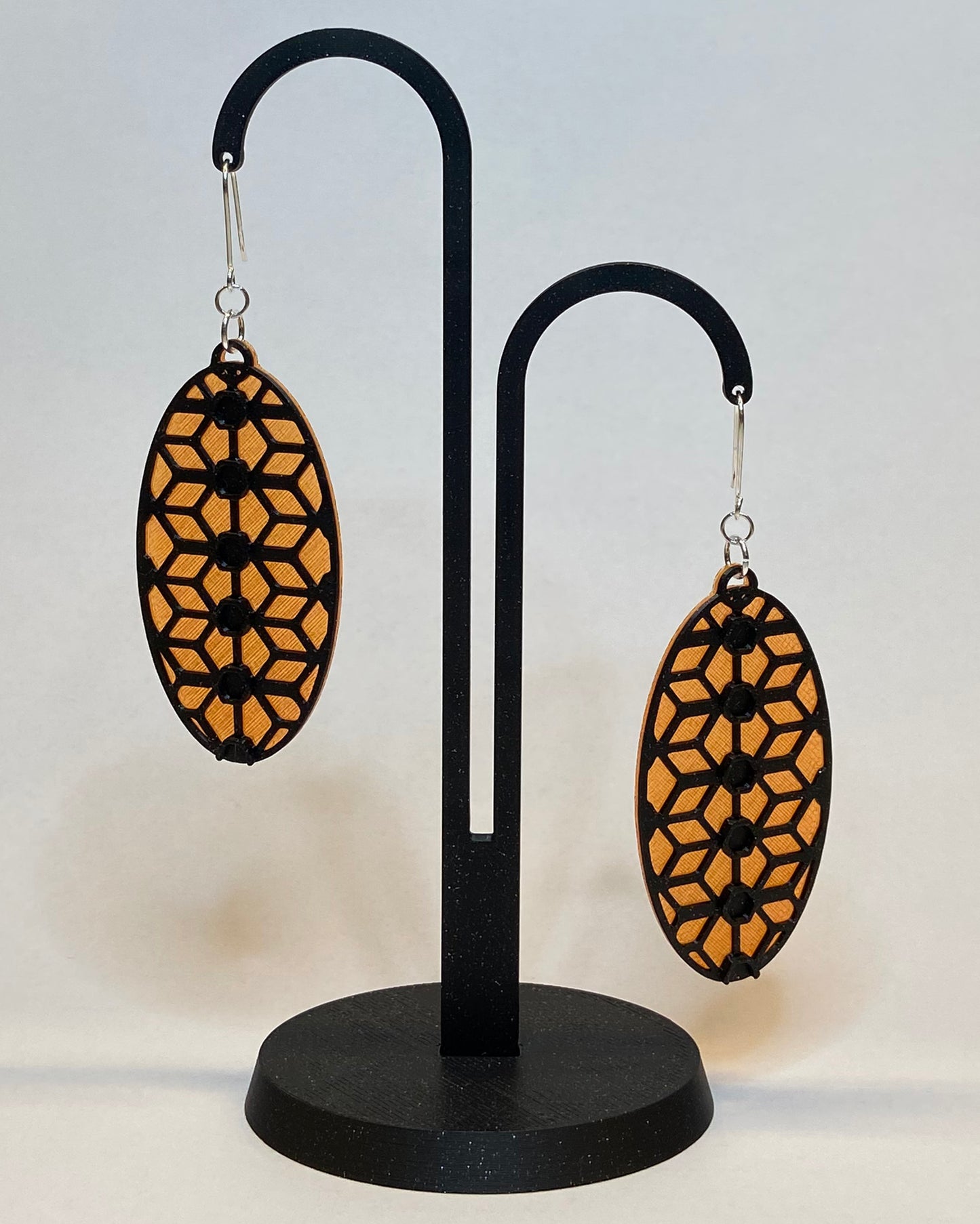 A pair of 3D-printed earrings with a lace inspiered design on a black stand against a light background. The earrings has two colours, an orange coloured bottom and contrasting black pattern. The fish hook is made of silver.
