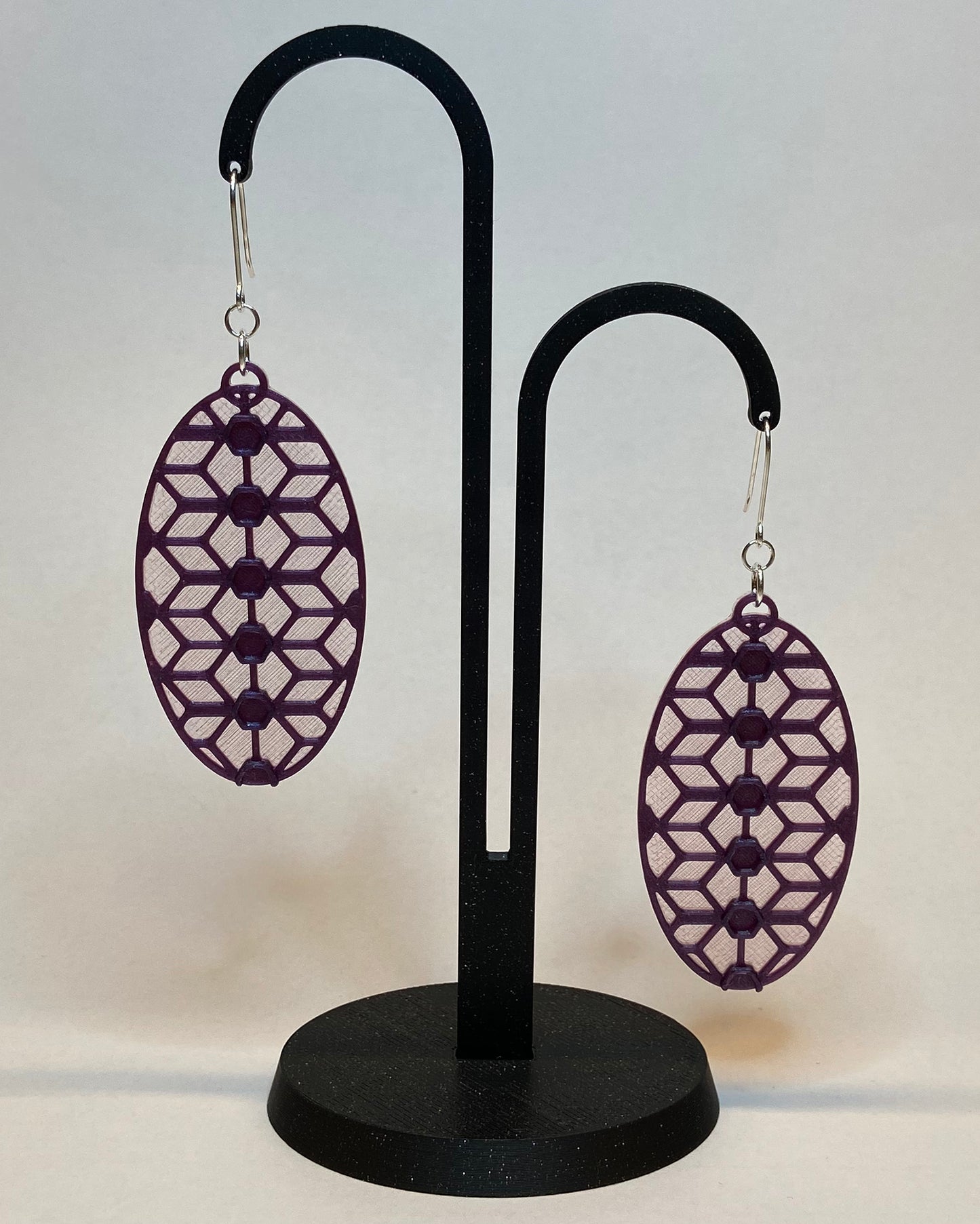 A pair of 3D-printed earrings with a lace inspiered design on a black stand against a light background. The earrings has two colours, a pink coloured bottom and contrasting purple pattern. The fish hook is made of silver.