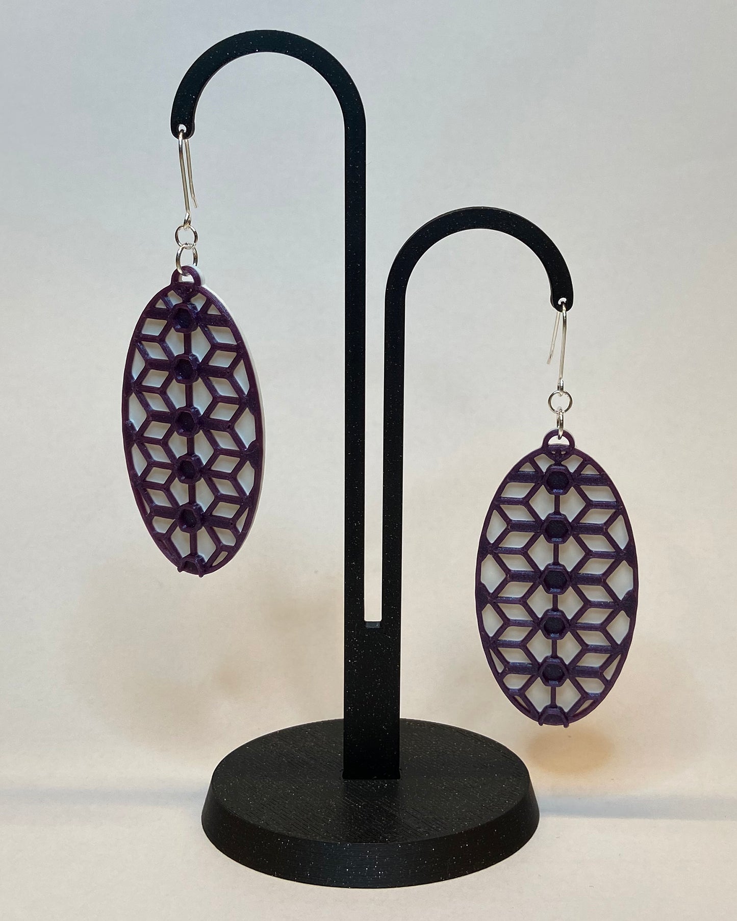 A pair of 3D-printed earrings with a lace inspiered design on a black stand against a light background. The earrings has two colours, a white coloured bottom and contrasting purple pattern. The fish hook is made of silver.