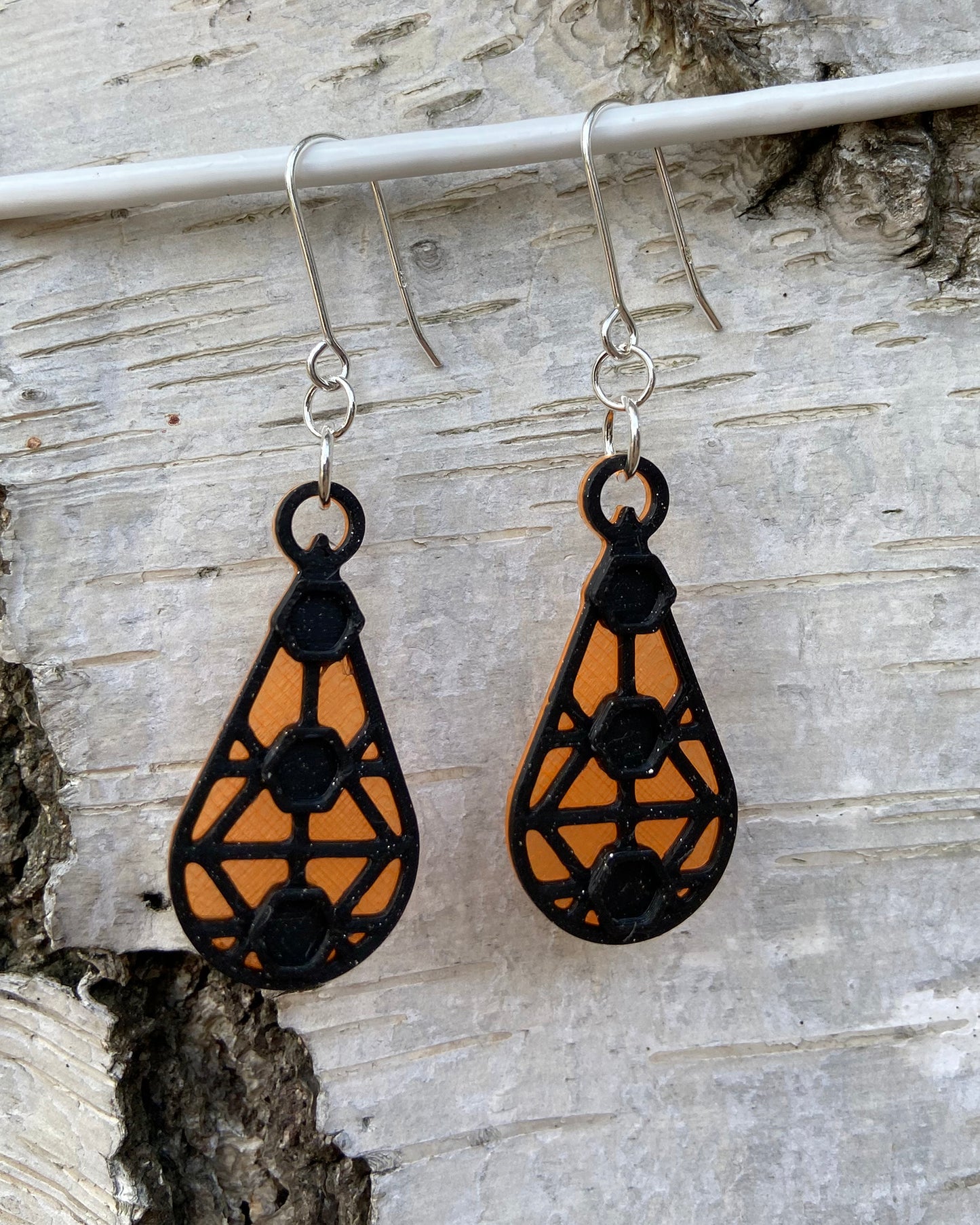 Closeup of a pair of orange and black geometric earrings, haning on a wire on a birchtree.