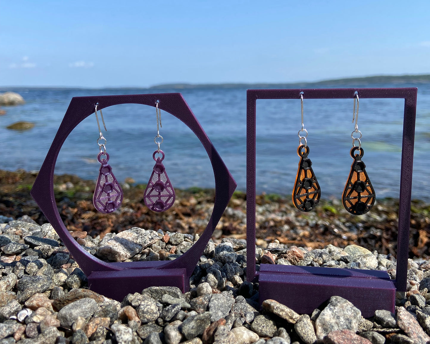 Two pairs of earrings, displayed on stands against a rocky beach and the sea and a blue sky. The earrings to the left are light pink and purple and the earrings to the right are orange and black.