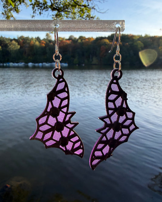 A pair of dangling earrings with a bat-shaped silhouette and a purple & black stained-glass like pattern, hanging from a clear stand against a blurred outdoor background of water and foliage.