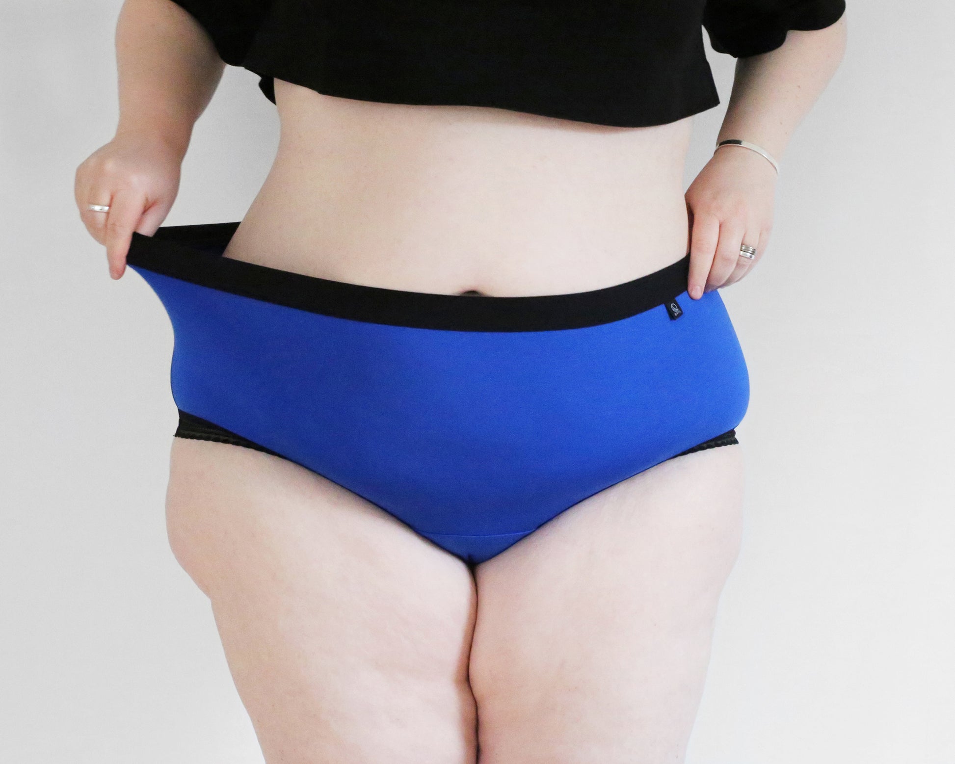 Front view of model wearing The Bamboo Hipster - Limited Edition in blue colour and black trims, stretching out the garment waist with one hand and the other hand on her hip.