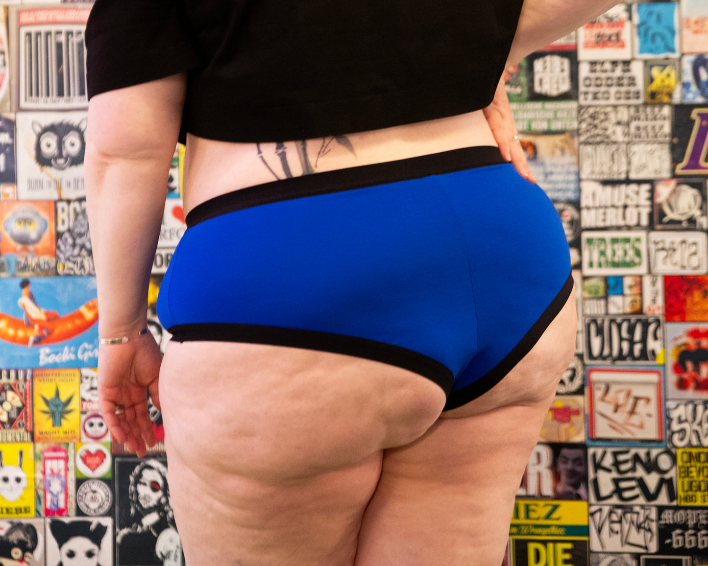 Back view of a model wearing a pair of Bamboo Brazilians in the blue colourway, also wearing a black t-shirt. She is standing infront of a wall with graphic designs, in a realxed pose with one hand on her hip.