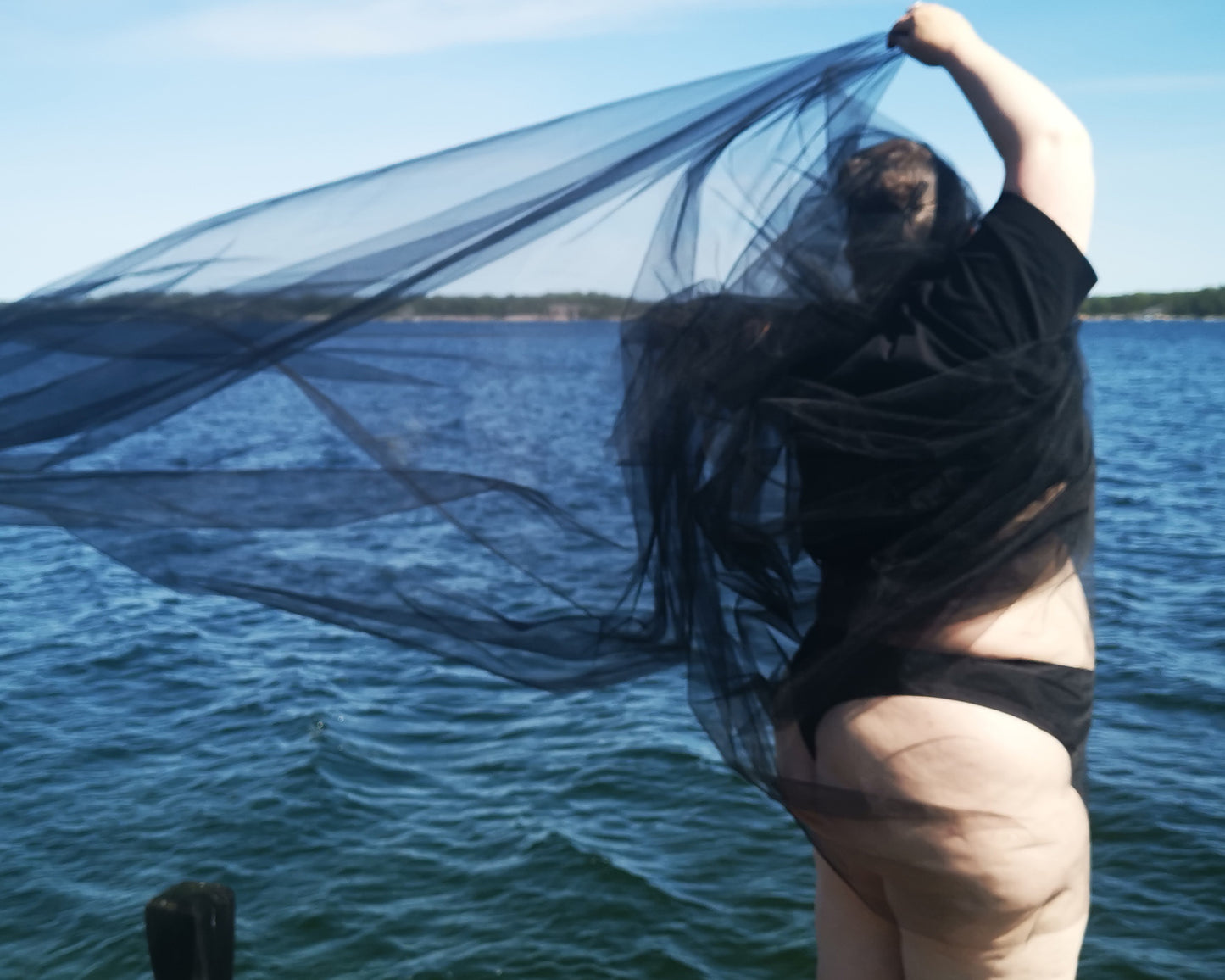 A model is wearing a pair of The Bamboo Thong in the black colourway. She is standing in front the sea, with a a sheer black fabric draped over hern front of a body of water shoulders. The fabric is flowing and billowing in the wind.