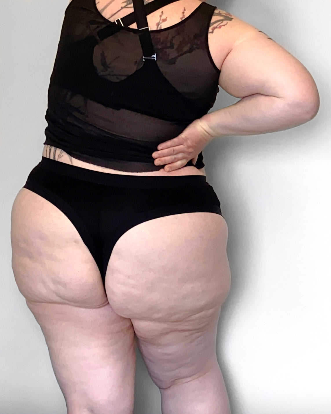 Back view of a model wearing The Bamboo Thong in the black colourway, with a sheer top and bra. She is streching one arm above her head and holding her other hand at her waist, infront of a light background.