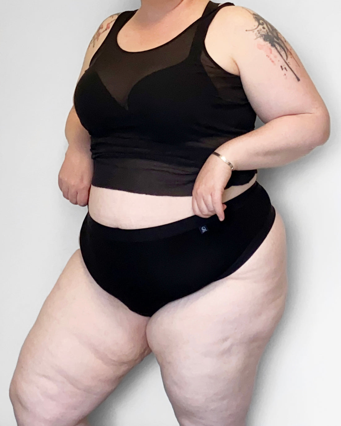 Frontside  view of a model wearing The Bamboo Thong in the black colourway, with a sheer top and bra. She is pulling at the waist of her knickers, infront of a light background.