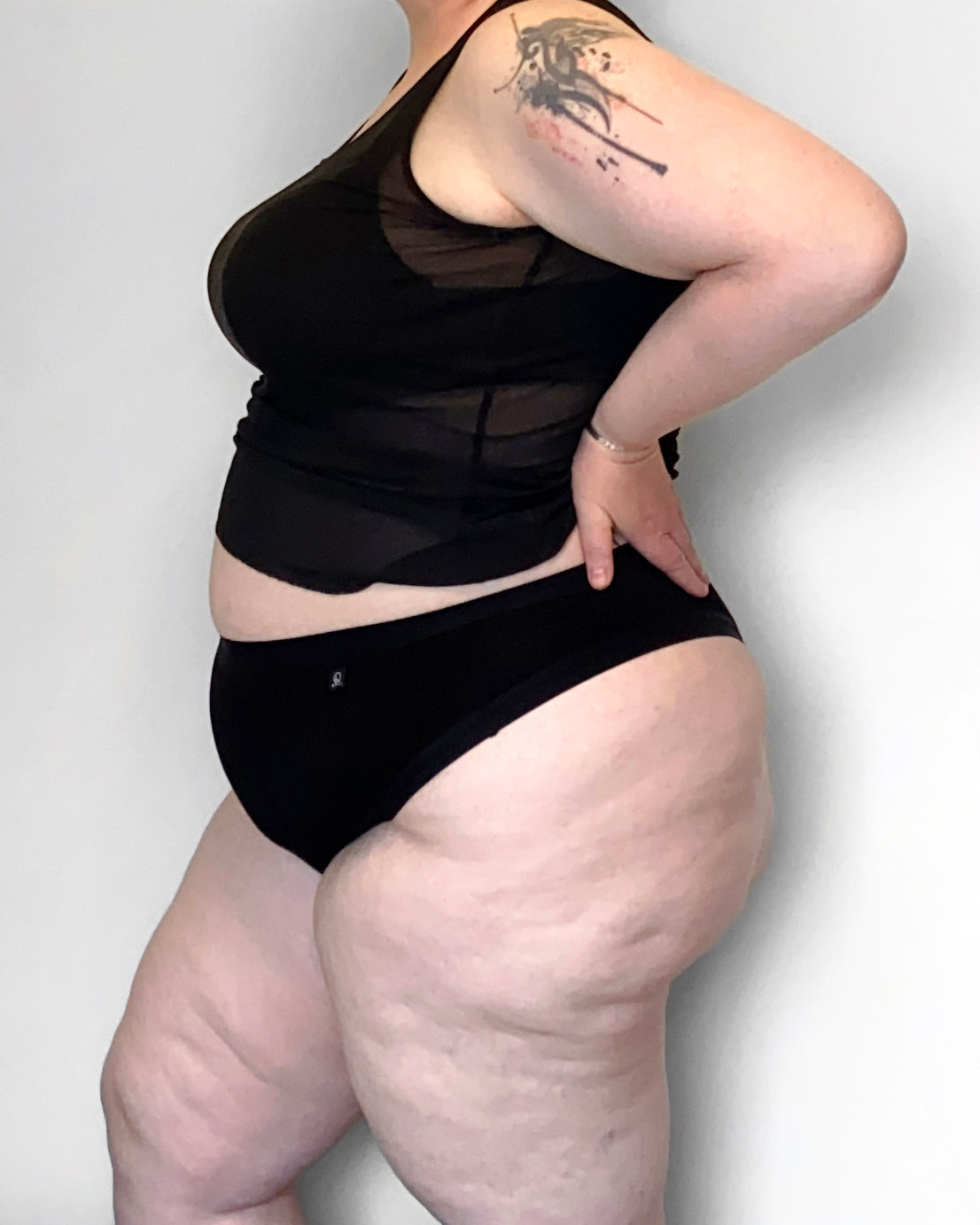Side view of a model wearing The Bamboo Thong in the black colourway, with a sheer top and bra. She is resting her hands on her small back, elbows turned back, infront of a light background.