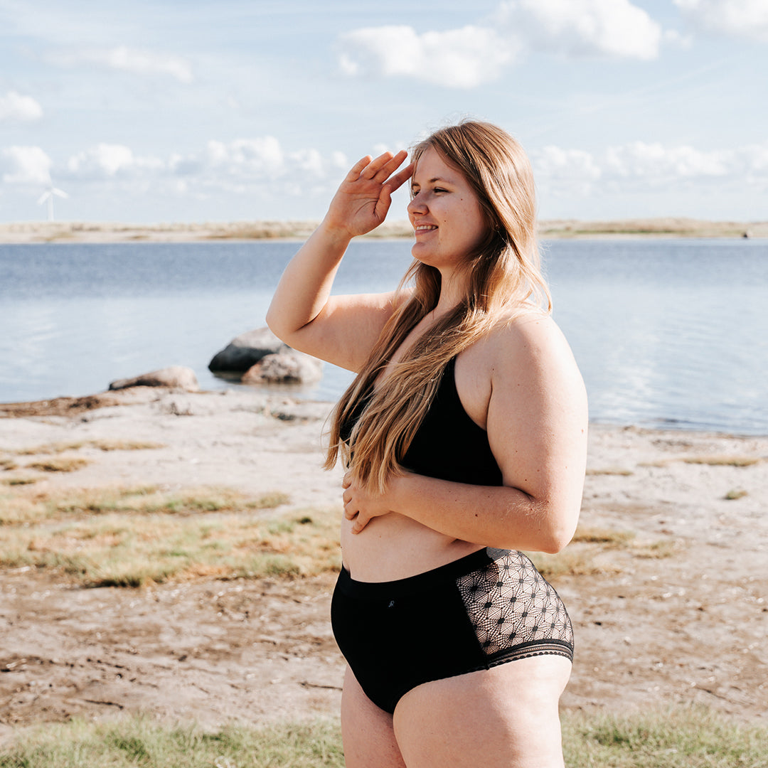 Model half turned towards viewer, arranging her hair with one hand, wearing The Mix Hipster. Water in the background. 