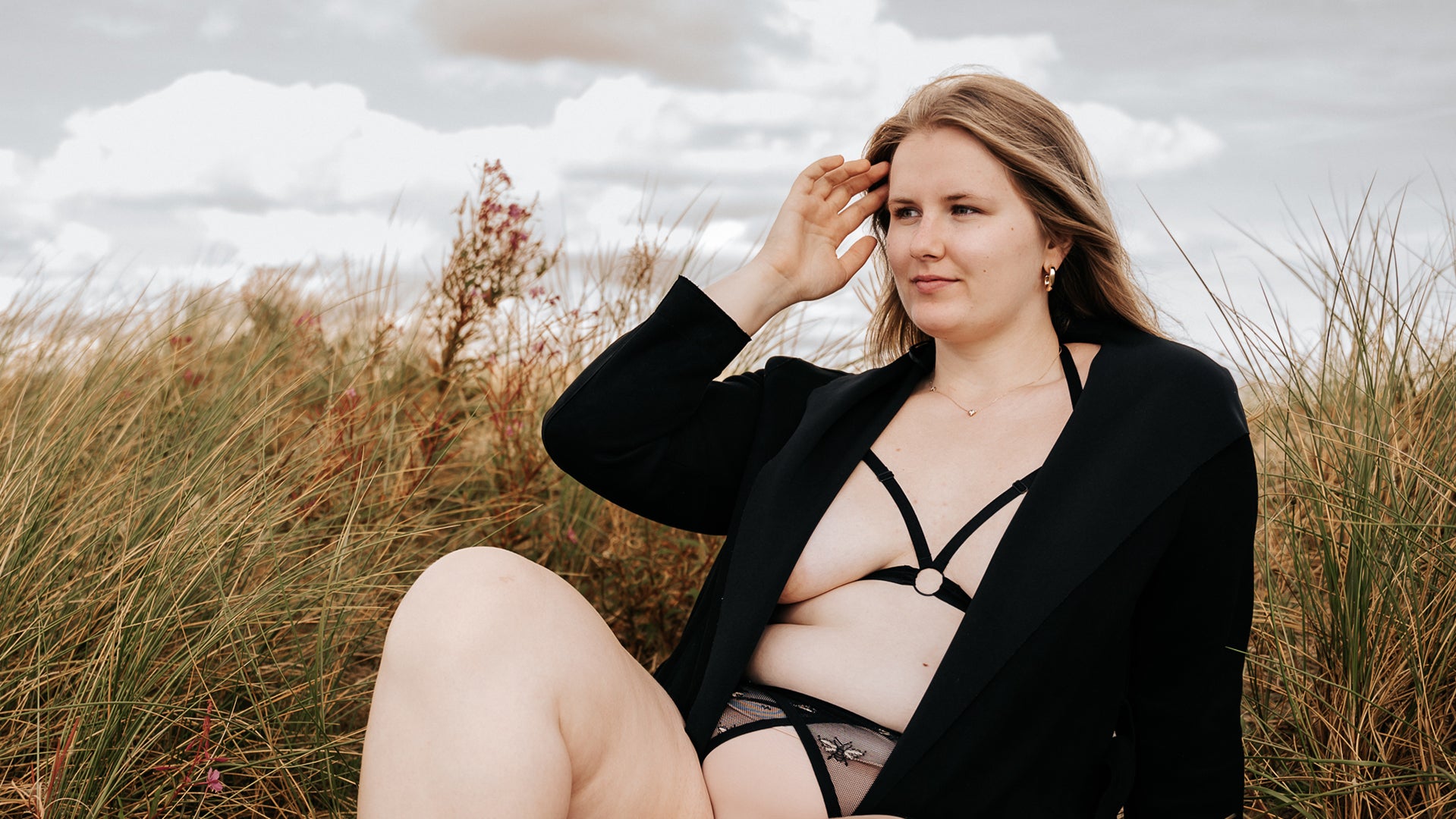 Smiling model wearing The Cage Bra under a jacket, relaxing in the grassy sand dunes facing slightly away from the viewer.