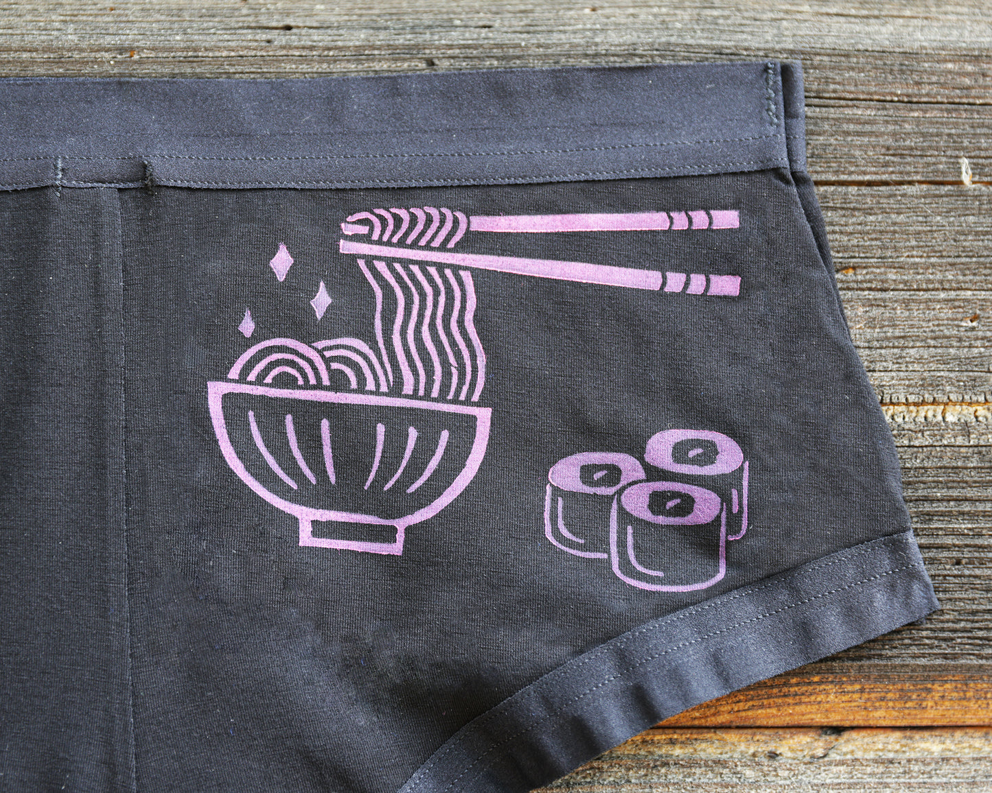 Black underwear laid flat on a rustic wooden surface, featuring a magenta coloured graphic of a bowl of ramen with chopsticks lifting noodles and three maki rolls.