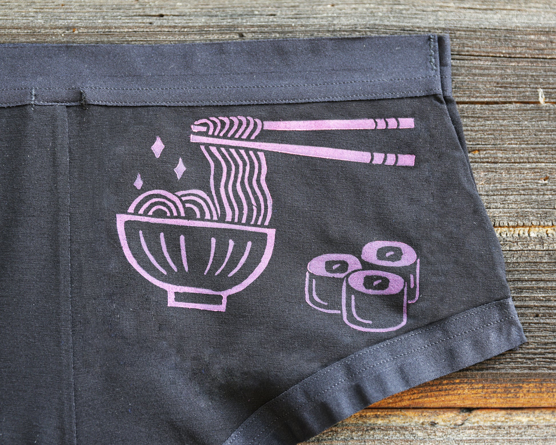 Black underwear laid flat on a rustic wooden surface, featuring a magenta coloured graphic of a bowl of ramen with chopsticks lifting noodles and three maki rolls.