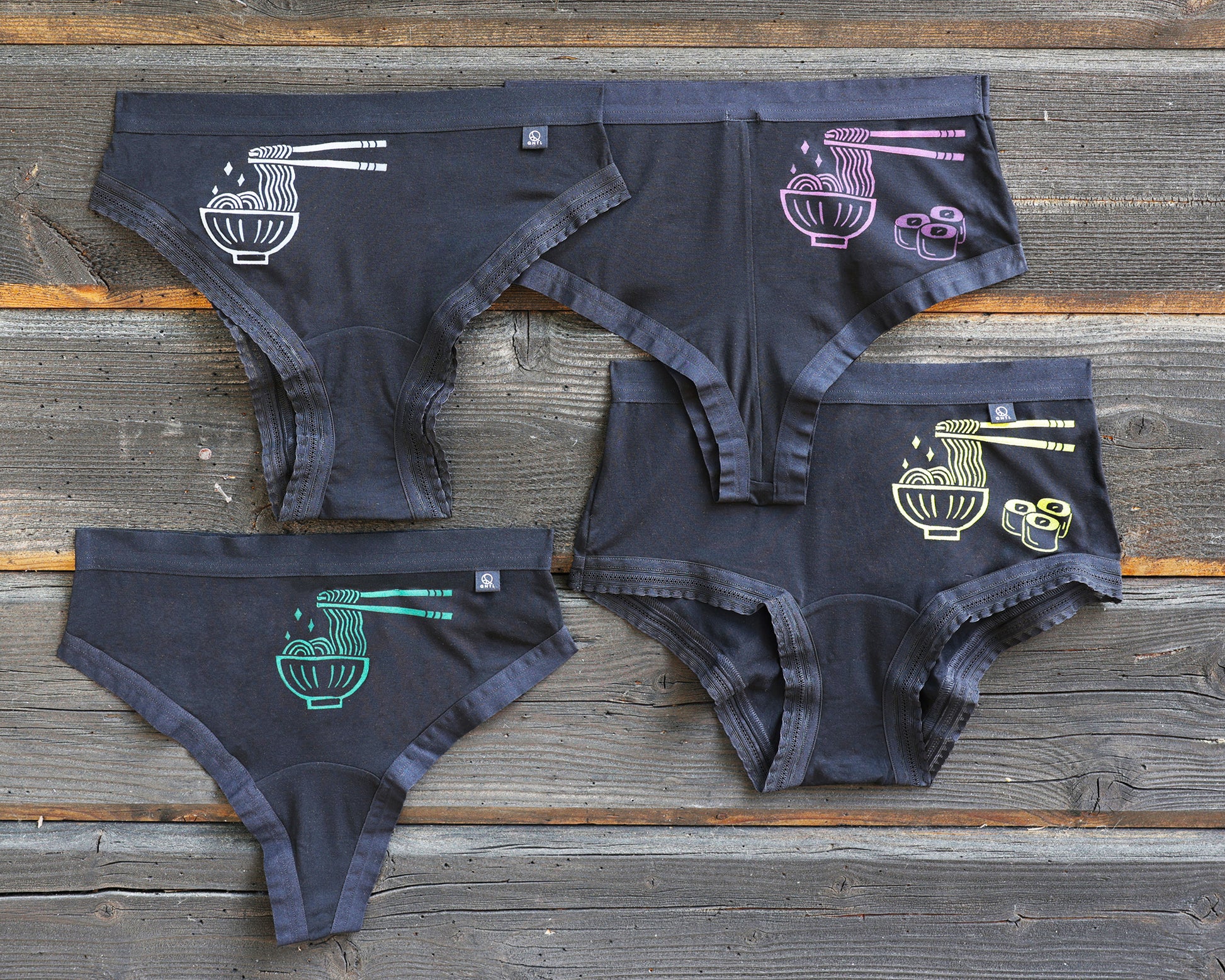 Four pairs of underwear on a rustic wooden plank background, each featuring a different colored graphic of a bowl of ramen with chopsticks and noodles; white, magenta, green and yellow. This is The Bowl of Ramen print!  The magenta and yellow prints alos has three small maki next to the bowl of ramen. 