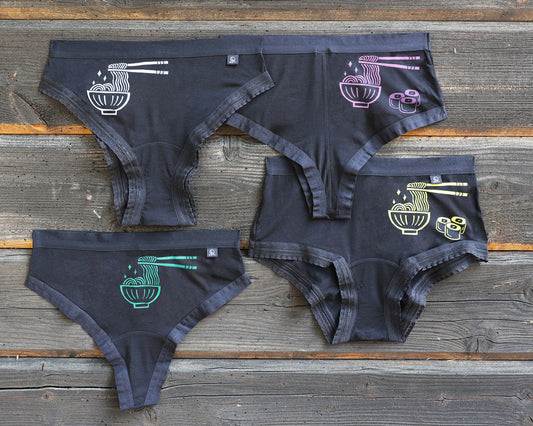 Four pairs of underwear on a rustic wooden plank background, each featuring a different colored graphic of a bowl of ramen with chopsticks and noodles; white, magenta, green and yellow. This is The Bowl of Ramen print!  The magenta and yellow prints alos has three small maki next to the bowl of ramen. 