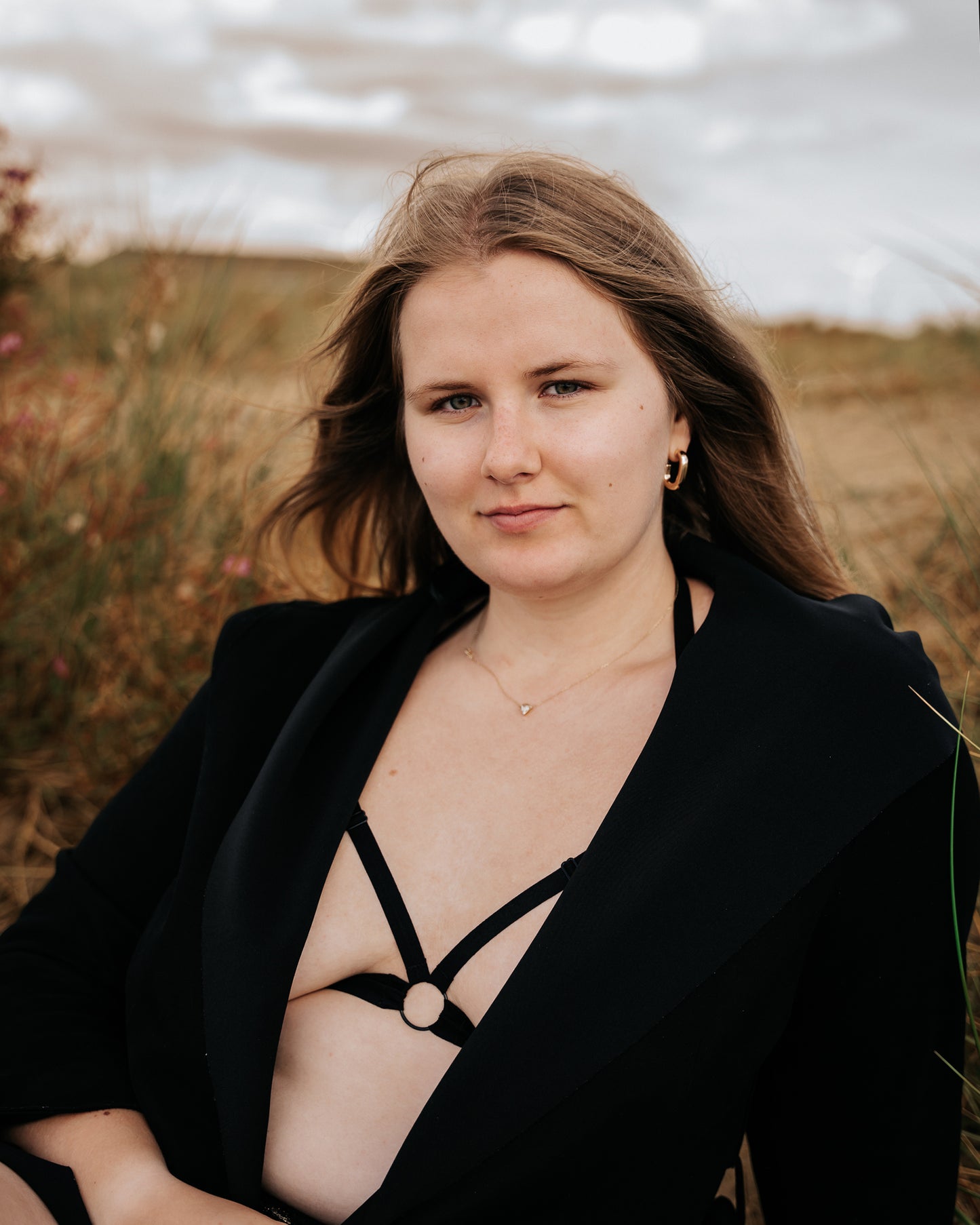 Front view of a smiling model wearing The Cage Bra under a jacket, relaxing in the grassy sand dunes looking directly at the viewer.