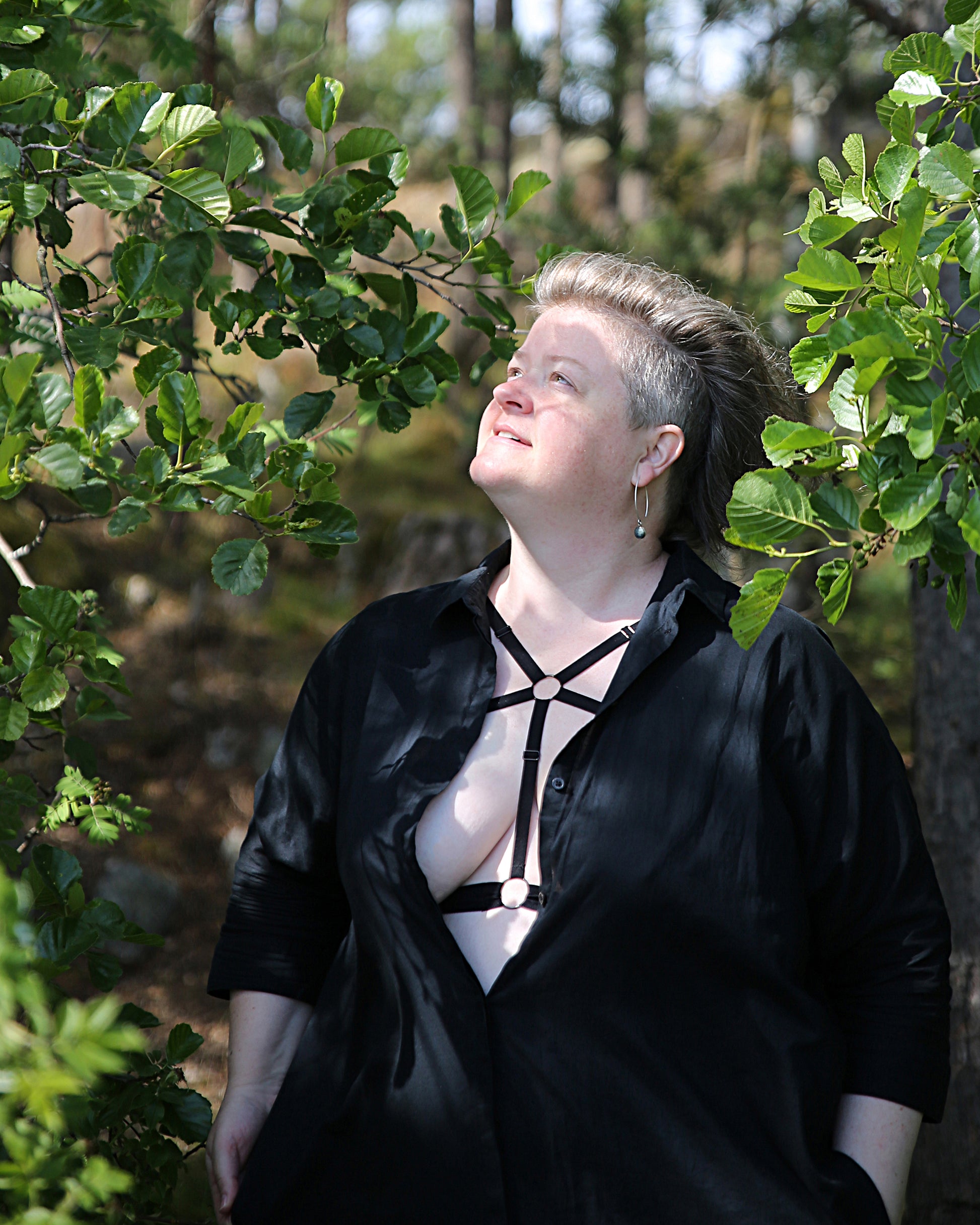 Front view of a smiling model looking up at the sun wearing The Cage Bra - Variation 1 under a shirt. The model is standing in a clearing, the trees are creating a shadowy pattern across her body.