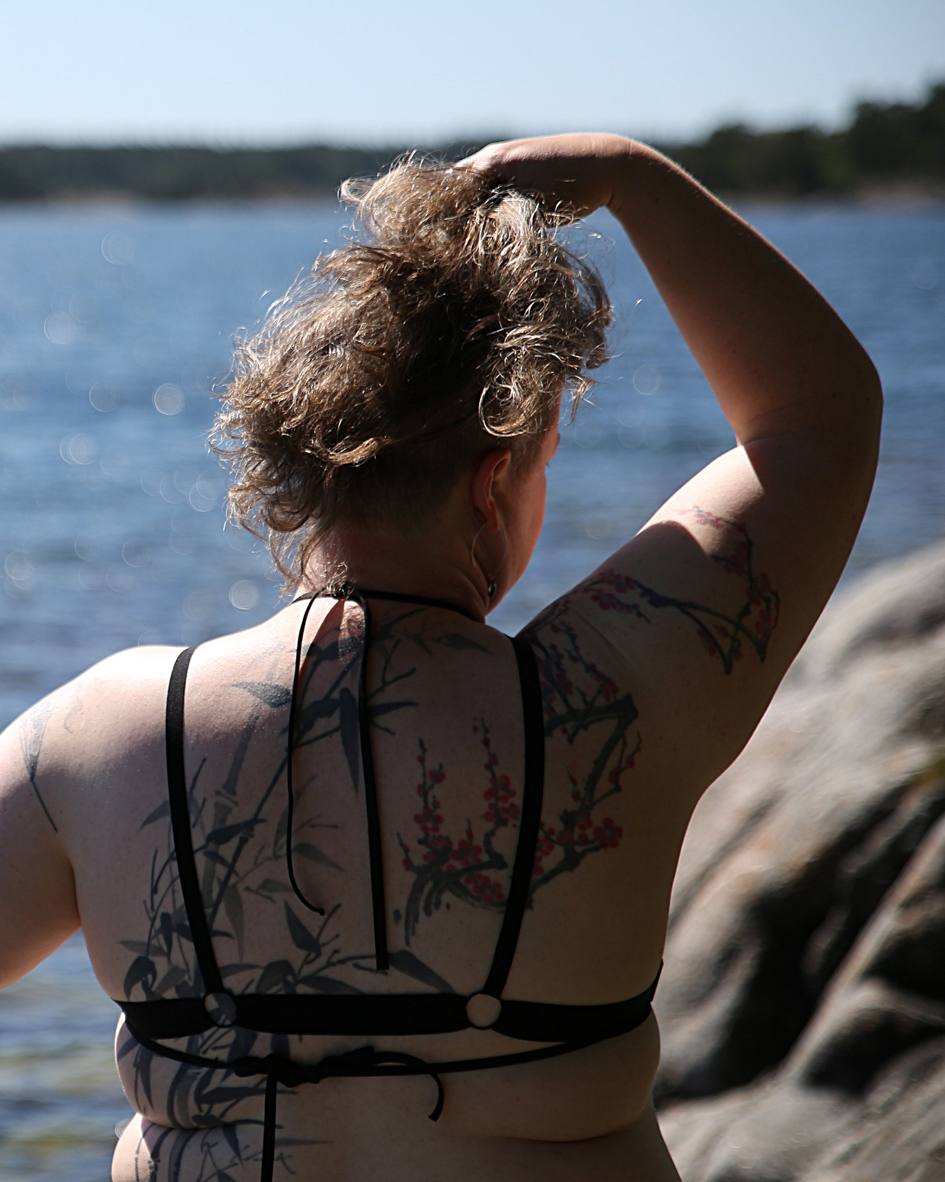 Back view of a model wearing The Cage Bra - Variation 1 over a bikini. The model is standing in front of a cliff and the sea, one hand in her hair.