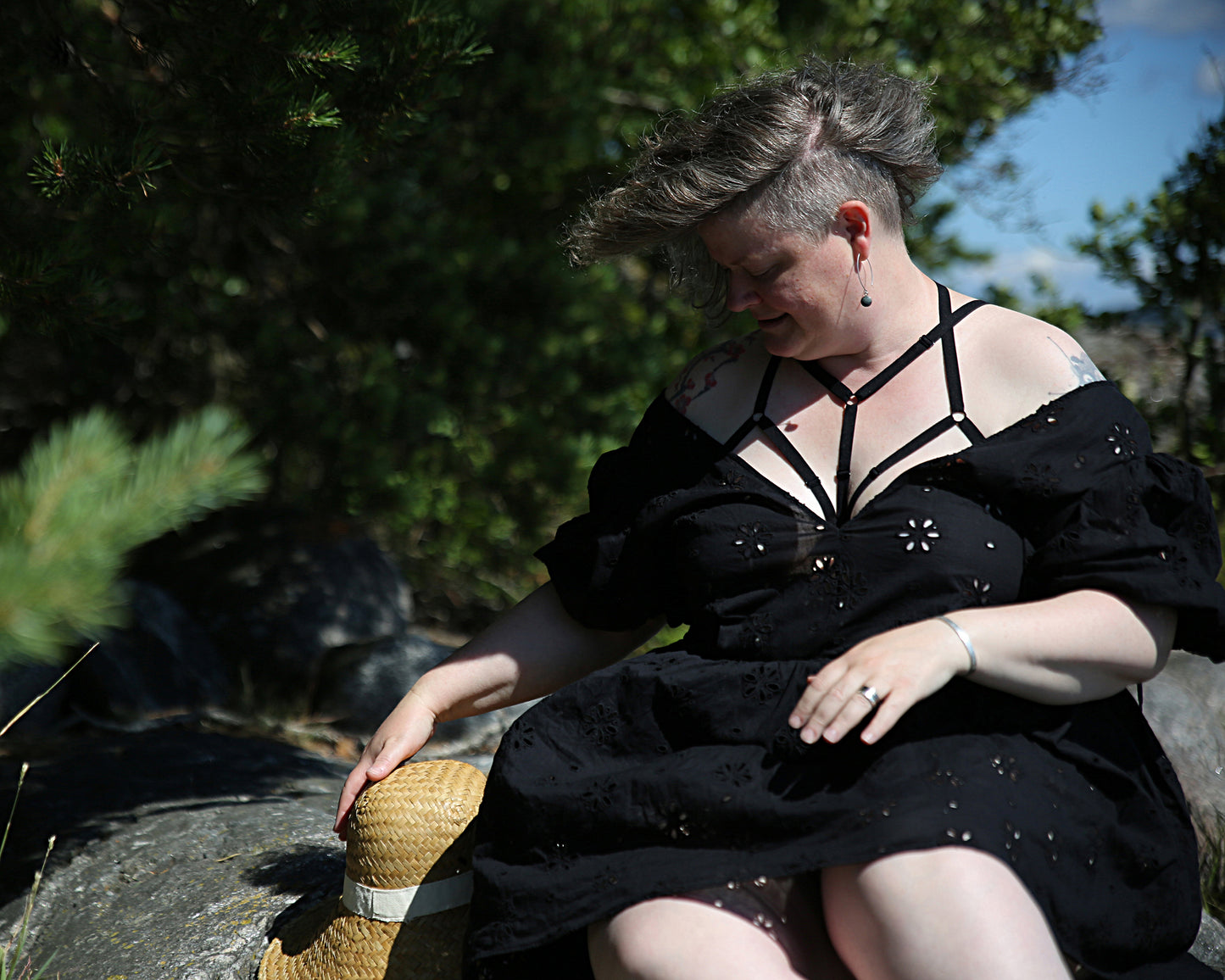 Front view of a smiling model wearing The Cage Bra - Variation 2 under a dress, relaxing on a cliff with one hand on top of a straw hat that is next to her on the cliff she is sitting on. The model is looking down on the hat.