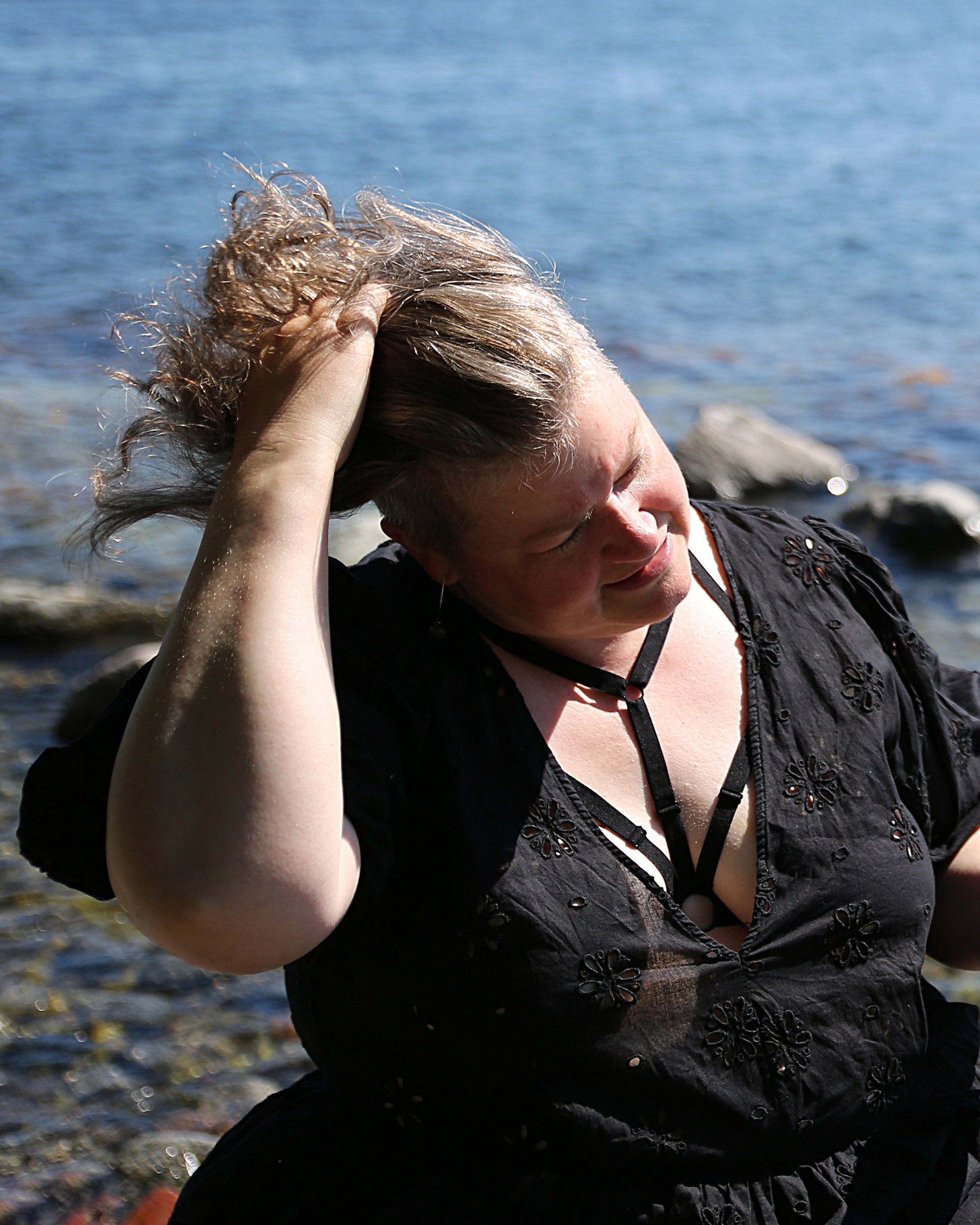 Front view of a smiling model wearing The Cage Bra - Variation 2 under a dress, arranging her hair with one hand and the back to the sea. 