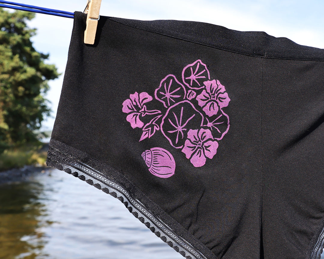Black underwear with a pink indian cresse and shell graphic, hanging from a clothesline outdoors near water.