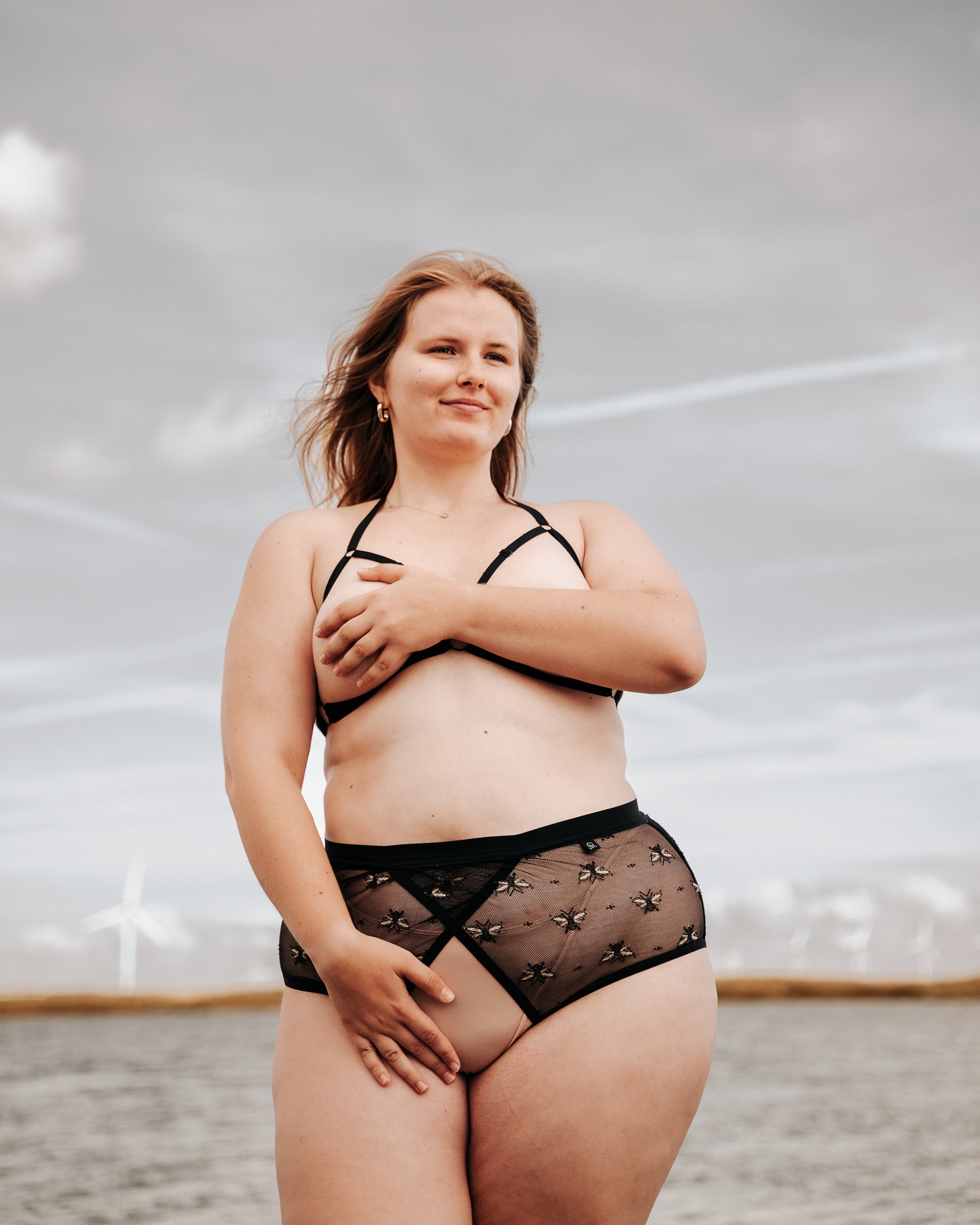 Front view of a smiling model wearing The Crossover Hipster and The Cage Bra, showing the overlapping lace panels of the knicker. The model is wearing a nude thong under The Crossover Hipster and her arm and hand is covering her nipples.