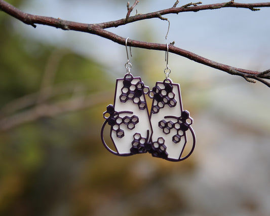 A pair of geometrical shaped earrings with a  honeycomb inspiered designe, haning on a branch. The earrings have a white coloured background and the pattern has a glittering purple colour.