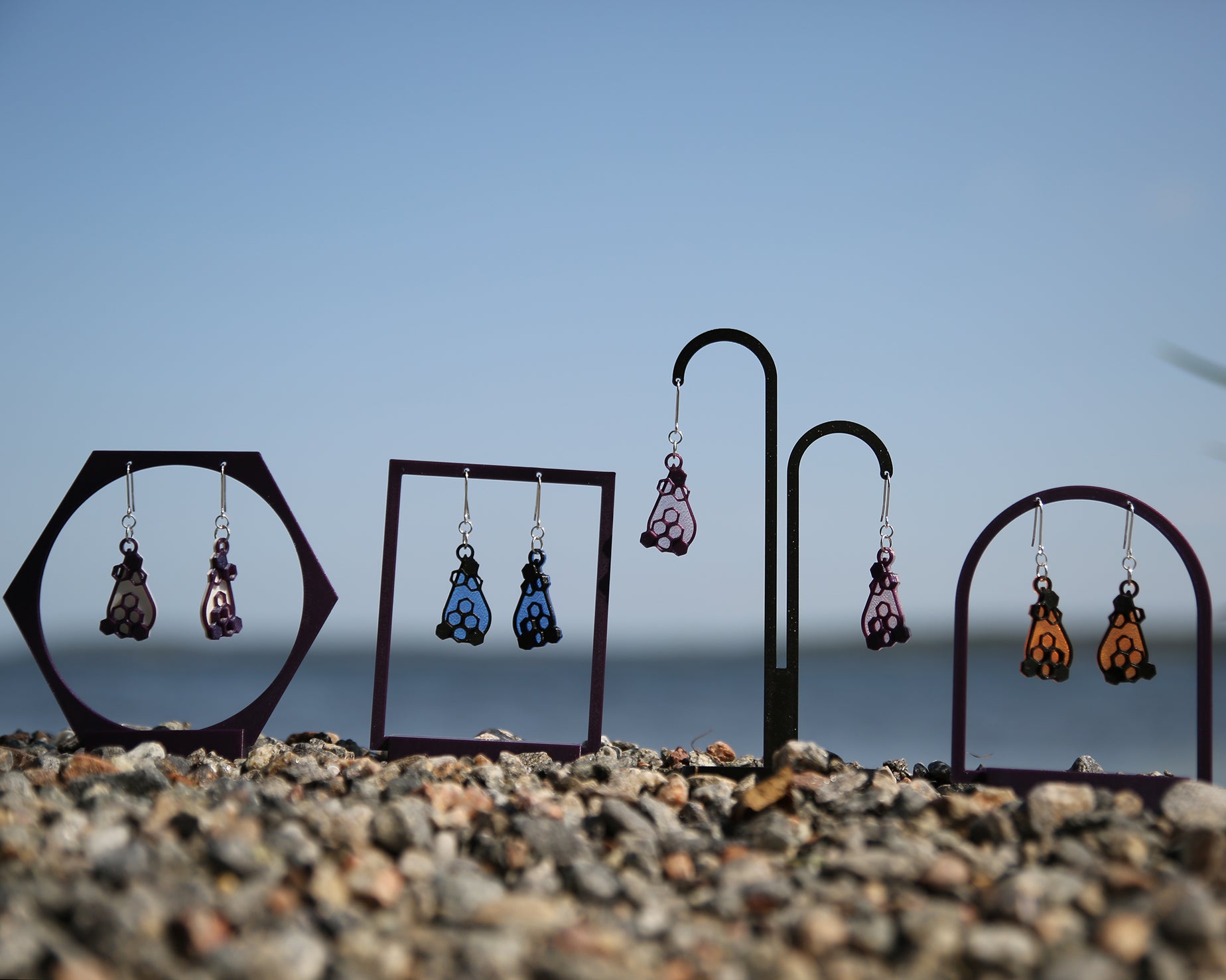 Colorful earrings with a honeycomb design, displayed on stands against a blue sky and rocky ground.