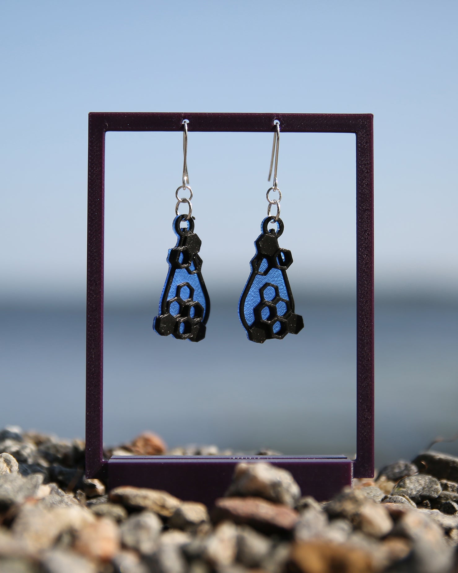 A pair of blue and black earrings with a honeycomb design, haning on a purple stand with a blurred natural background.